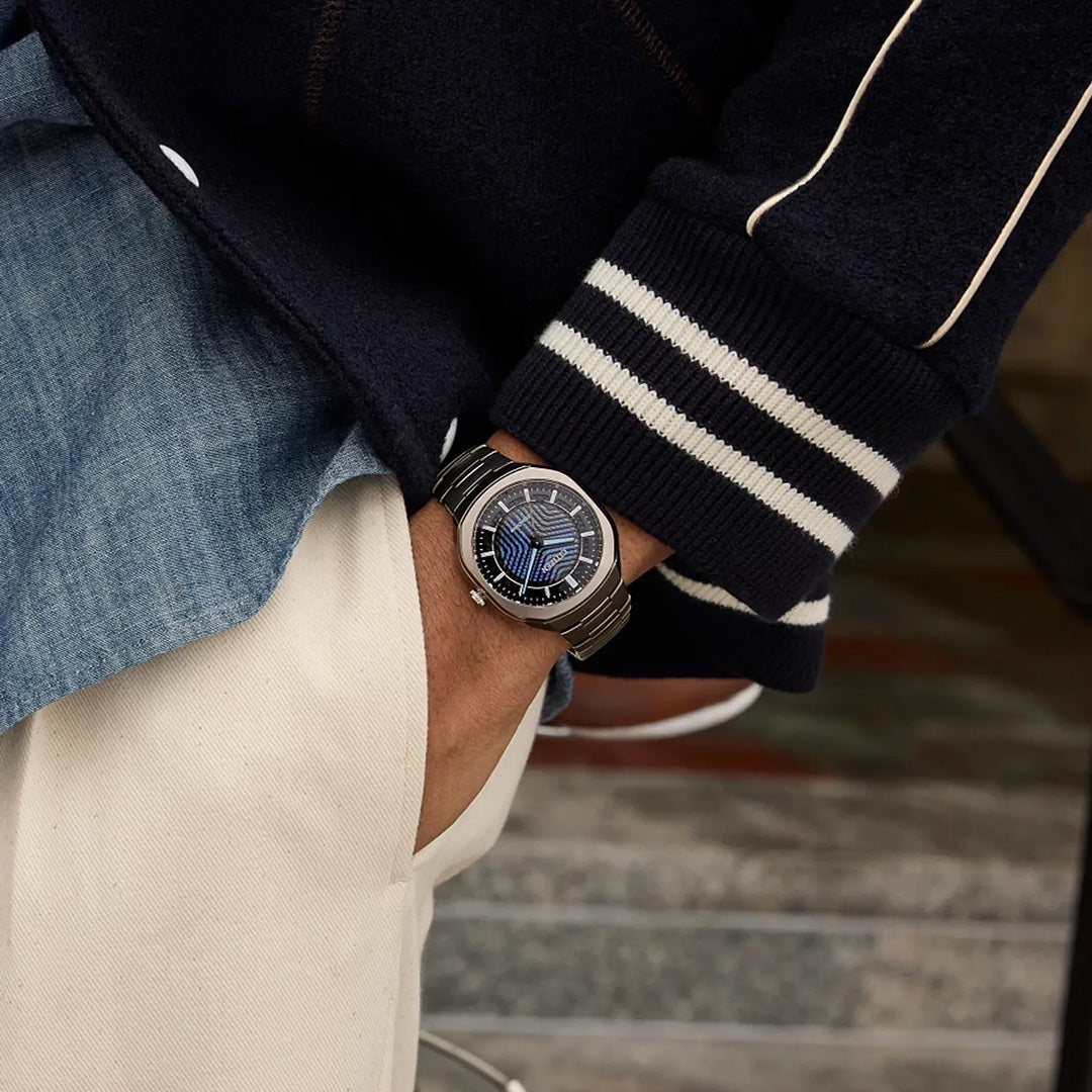 A person wearing a striped jacket and beige pants showcases the CITIZEN Photon Eco-Drive 39.6mm Watch Limited Edition of 5,000 Pieces, featuring a dark face and metallic Super Titanium band, standing near stairs—perfectly embodying CITIZEN’s fusion of sport luxury and modern elegance.