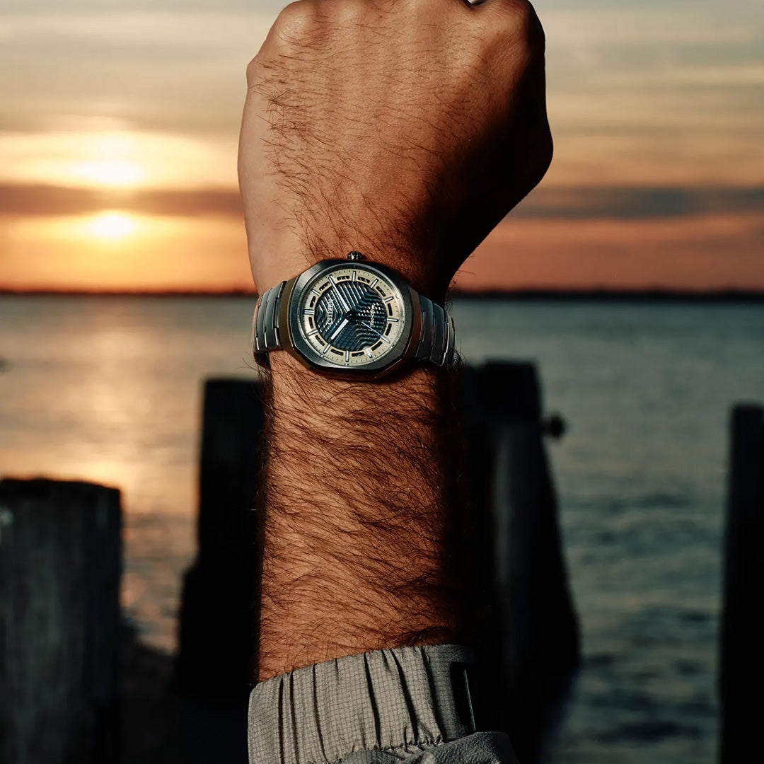 A raised arm wearing the CITIZEN Photon Eco-Drive 39.6mm Watch, Limited Edition of 5,000 pieces, is seen in front of a waterfront at sunset, with golden light reflecting on the water and wooden posts in the background.