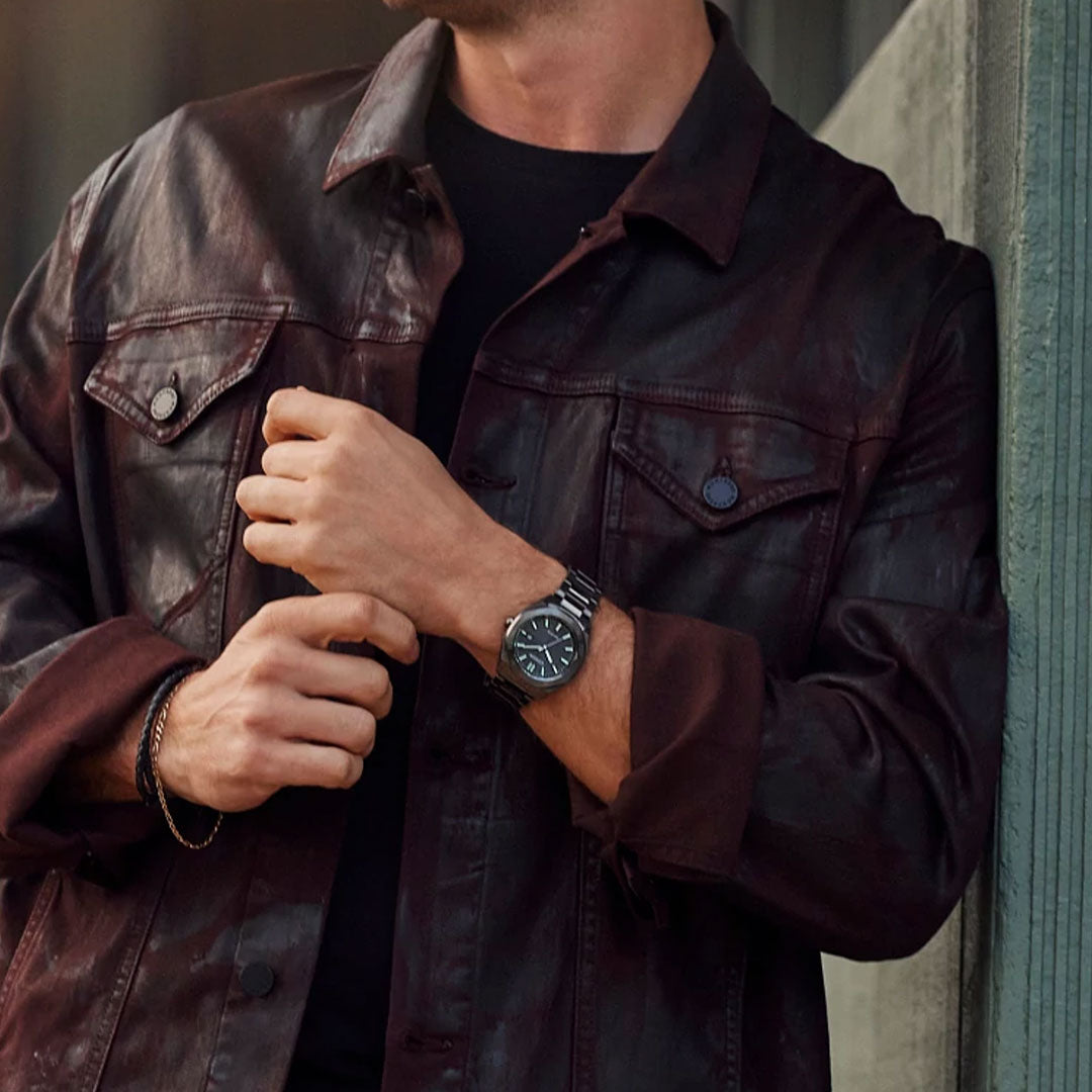 A person wearing a dark brown leather jacket adjusts the sleeve with their right hand. On their left wrist, they flaunt the CITIZEN Weekender Eco-Drive 41mm Watch, which features Eco-Drive technology and a stainless steel case. They stand near a wooden structure, partially visible from the neck down.