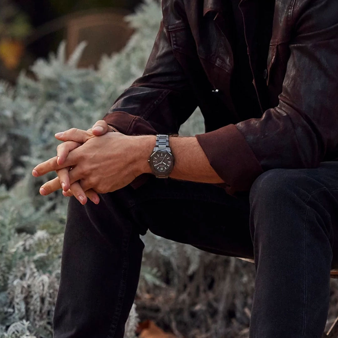 A person wearing a brown leather jacket and dark jeans is seated, hands clasped. They showcase the CITIZEN Weekender Eco-Drive 41mm Watch, which combines style with practicality. The lush green foliage in the background suggests an inviting outdoor setting.