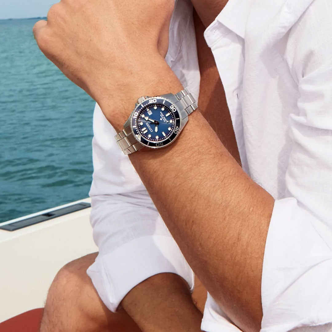Wearing a CITIZEN Promaster Dive Eco-Drive 41mm watch with a blue dial and bezel, someone sits on a boat in a white long-sleeve shirt by the water. This CITIZEN timepiece offers Eco-Drive technology and is water resistant to 200 meters.