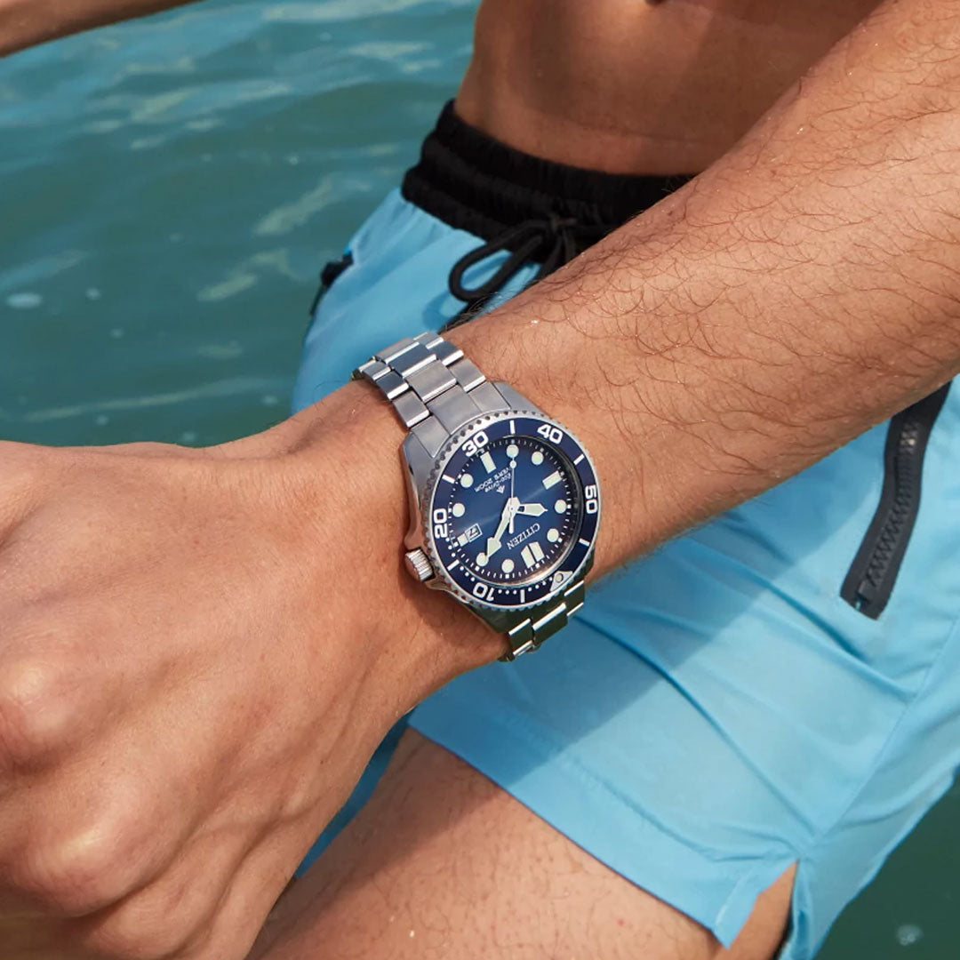 A person in blue swim trunks stands by water, displaying a CITIZEN Promaster Dive Eco-Drive 41mm Watch with a blue face, rotating bezel, and 200m water resistance on their wrist.