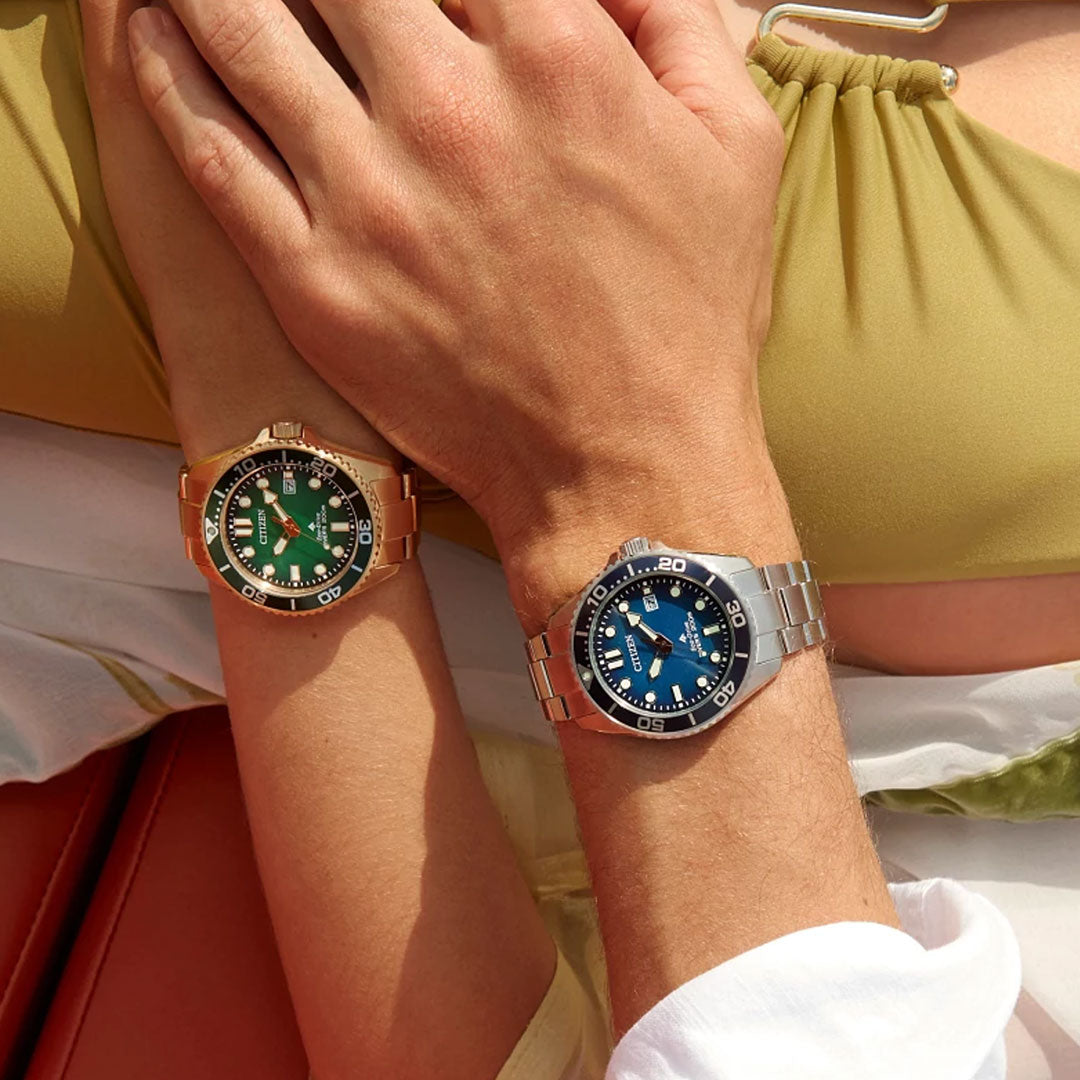 Two people’s arms rest together; one wears a CITIZEN Promaster Dive Eco-Drive 41mm Watch in gold with a green face, water resistant to 200 meters, while the other sports a silver watch with a blue face. Both are dressed in light summer clothing.