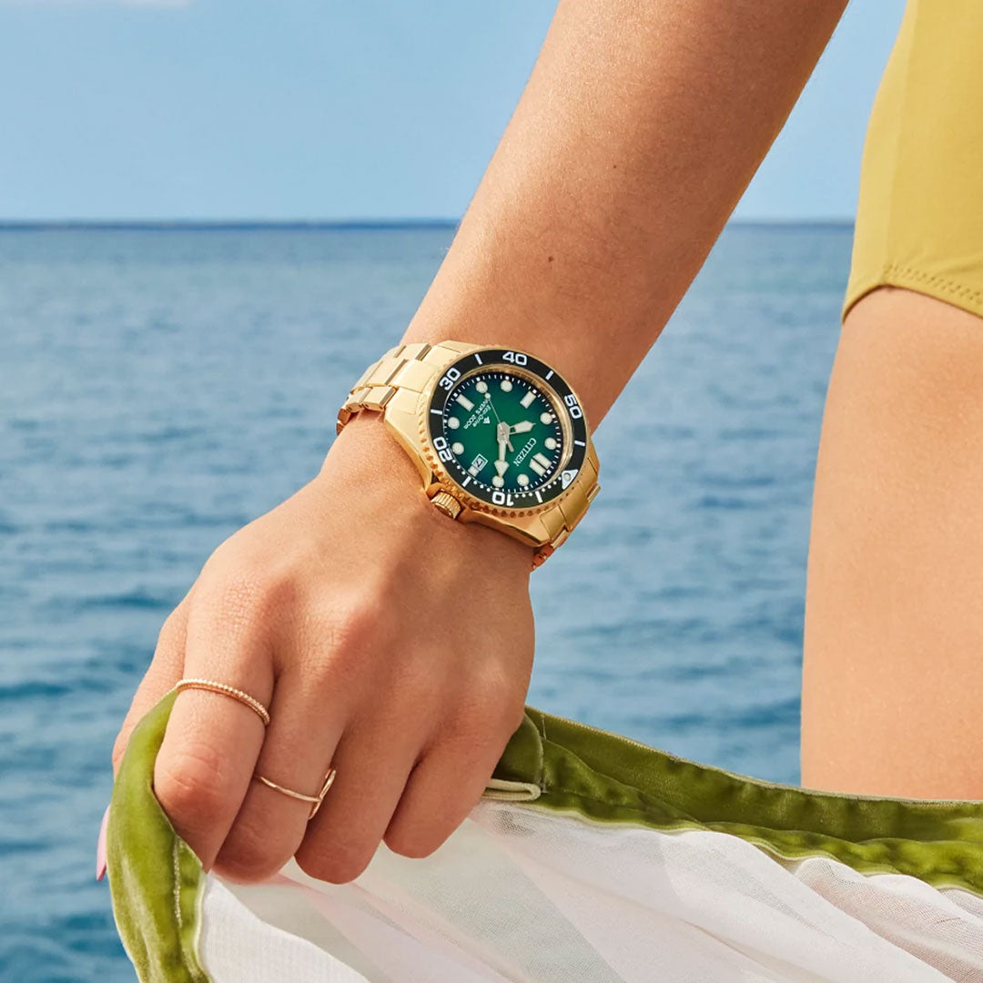 By the ocean, a person in a yellow swimsuit holds green and white fabric, wearing a CITIZEN Promaster Dive Eco-Drive 41mm Watch with a gold-tone stainless steel case and green dial. Blue water and sky create a serene backdrop.