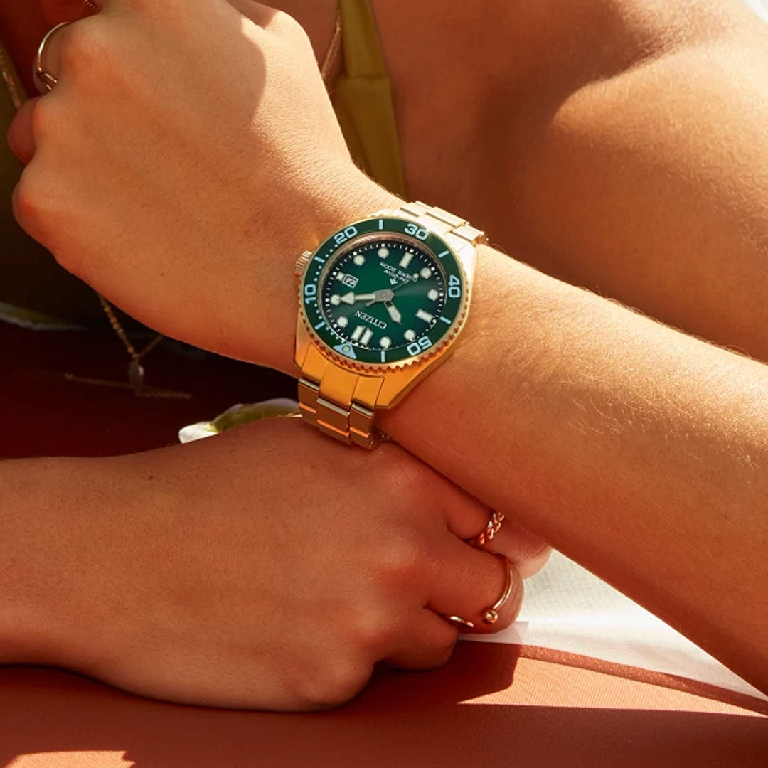A close-up shows a CITIZEN Promaster Dive Eco-Drive 41mm gold-tone stainless steel watch with a green face and white markings on a wrist, while the other hand, featuring a ring, rests gently above, partially revealing a sleeveless top.