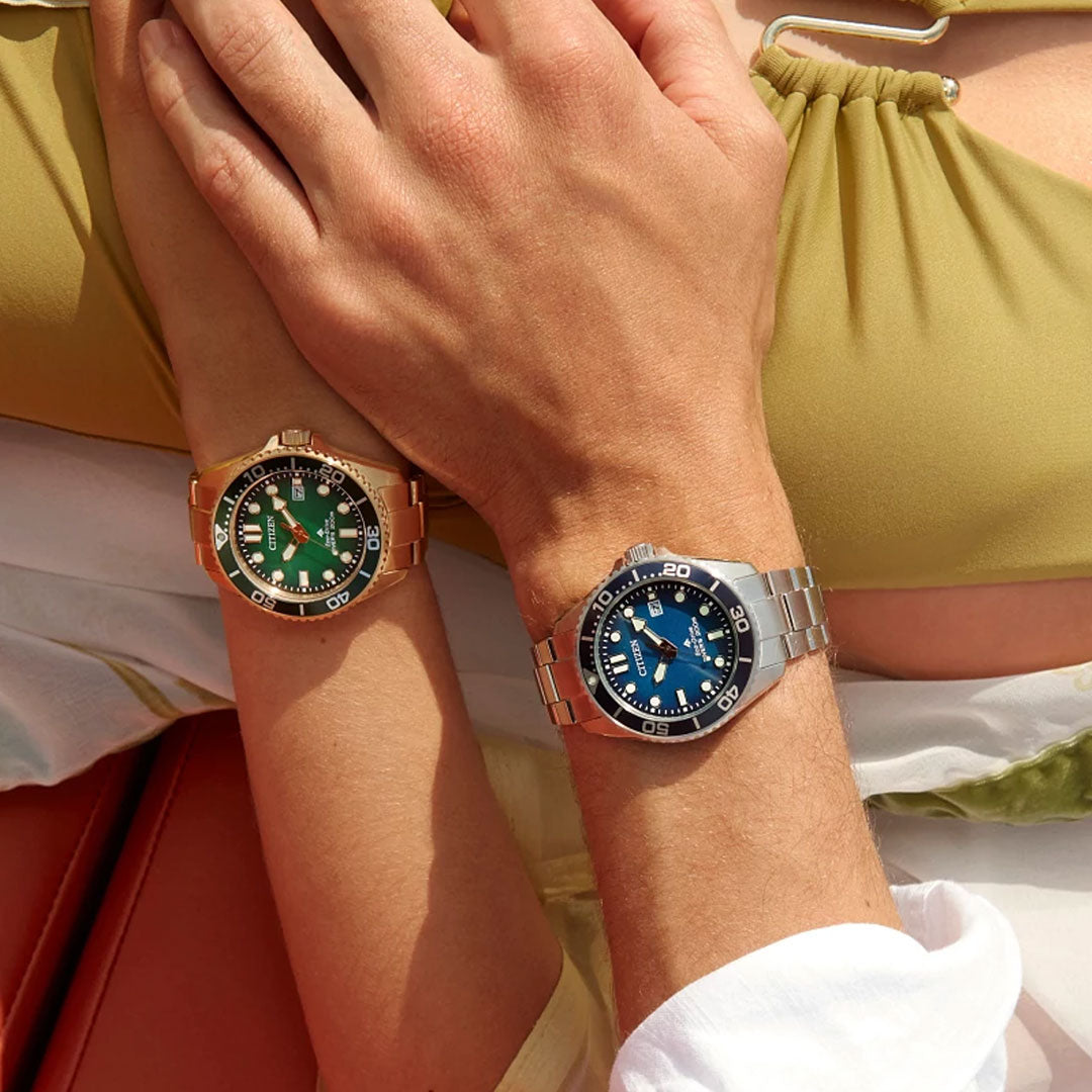 A close-up of two crossed arms shows a woman wearing the CITIZEN Promaster Dive Eco-Drive 41mm gold-tone watch, and a man sporting a silver watch with a blue face, both paired with light green and white outfits.