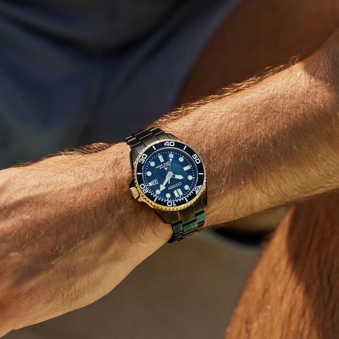 A close-up of a wrist wearing the CITIZEN Promaster Dive Light in Black Eco-Drive 40.6mm Watch, featuring a blue face, gold accents, and a black and gold metal strap. The arm rests on a knee in natural light.