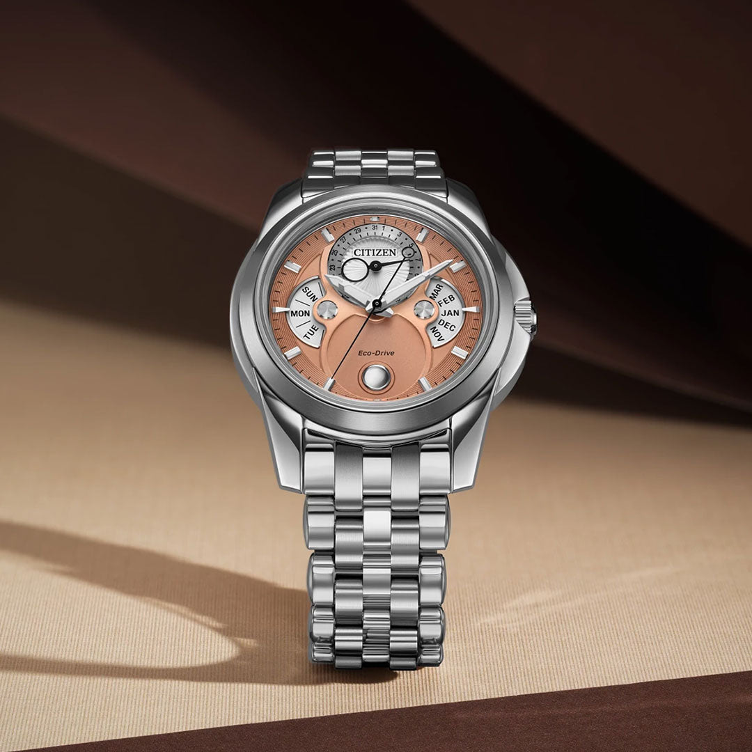 The CITIZEN Calendrier Eco-Drive 41.8mm Watch, silver with a stainless steel bracelet and copper-toned dial, features subdials for day, date, and month, shown on a beige and brown background.