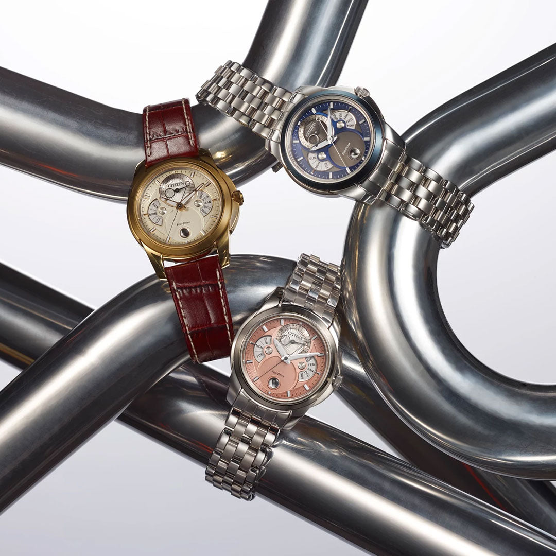 Three luxury wristwatches rest on metallic pipes: a gold case with brown leather strap, a CITIZEN Calendrier Eco-Drive 41.8mm watch in stainless steel with blue dial, and a silver case with salmon-pink dial and metal bracelet.