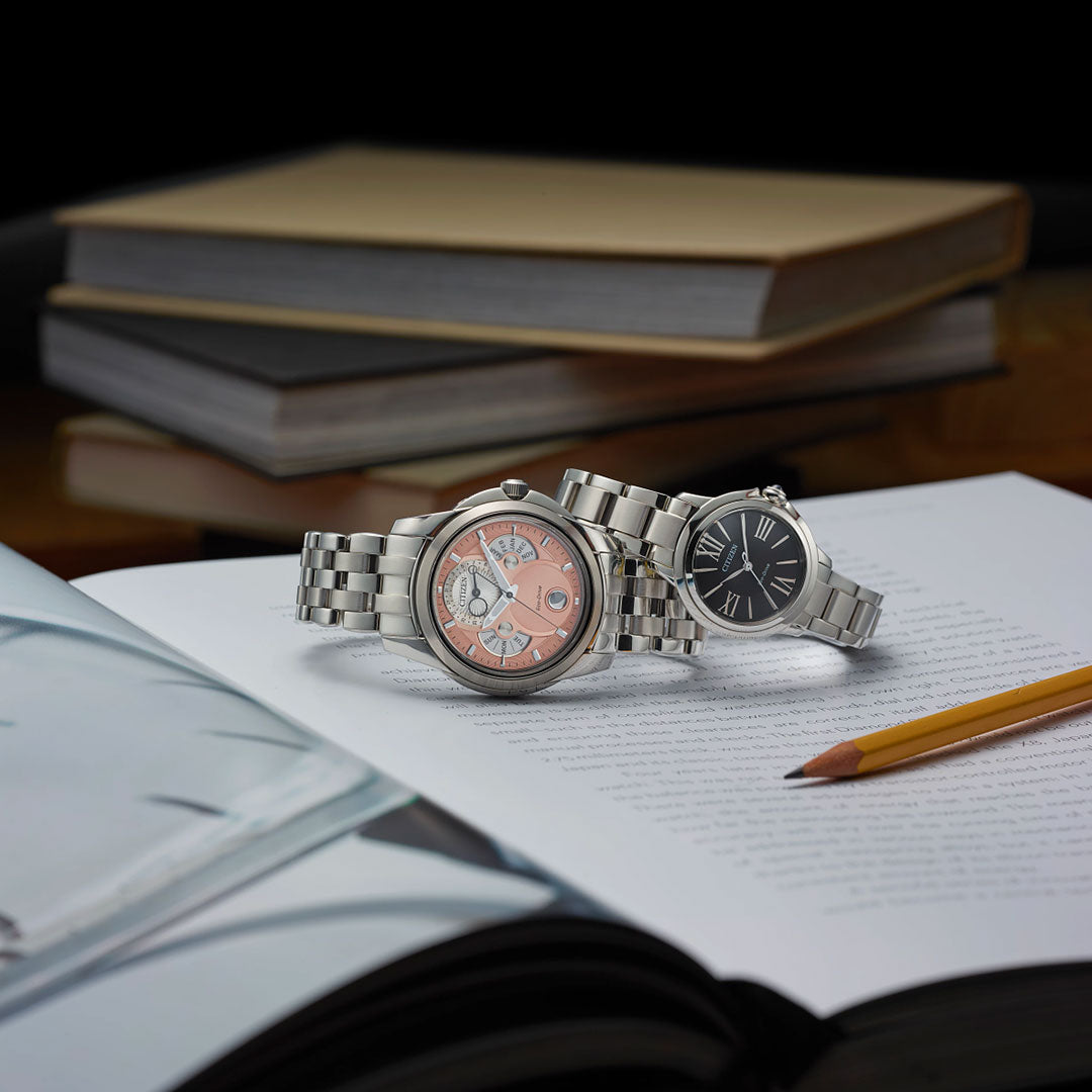 Two CITIZEN Calendrier Eco-Drive 41.8mm watches rest on an open book beside a yellow pencil, with stacked books in the background adding to the scholarly atmosphere.