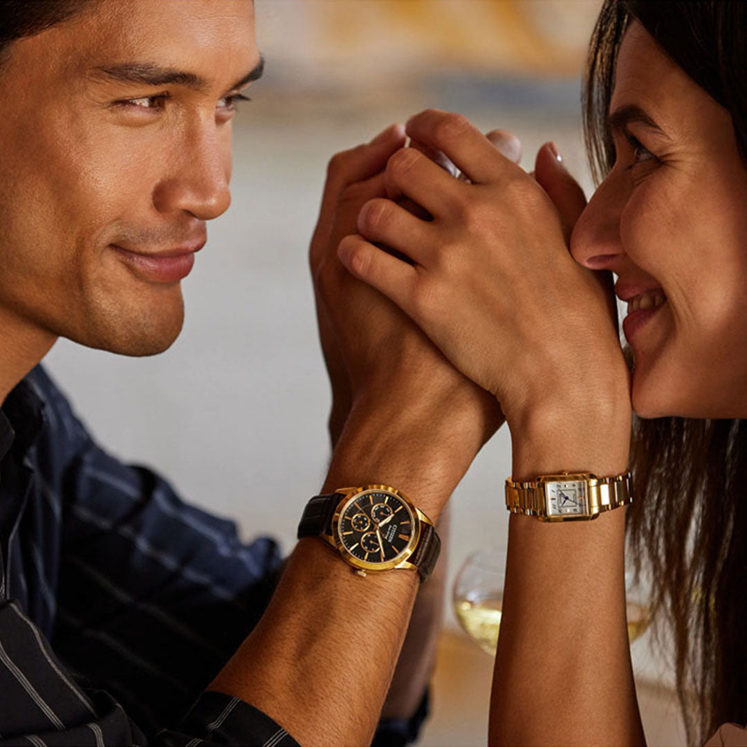 A man and a woman smile at each other while holding hands across a table. Both wear sophisticated timepieces; the man's CITIZEN Rolan Eco-Drive 40mm Watch, featuring a black and gold design with a leather strap, complements the woman's elegant gold, rectangular design. Two glasses of white wine are in the background, completing their vintage-inspired style.