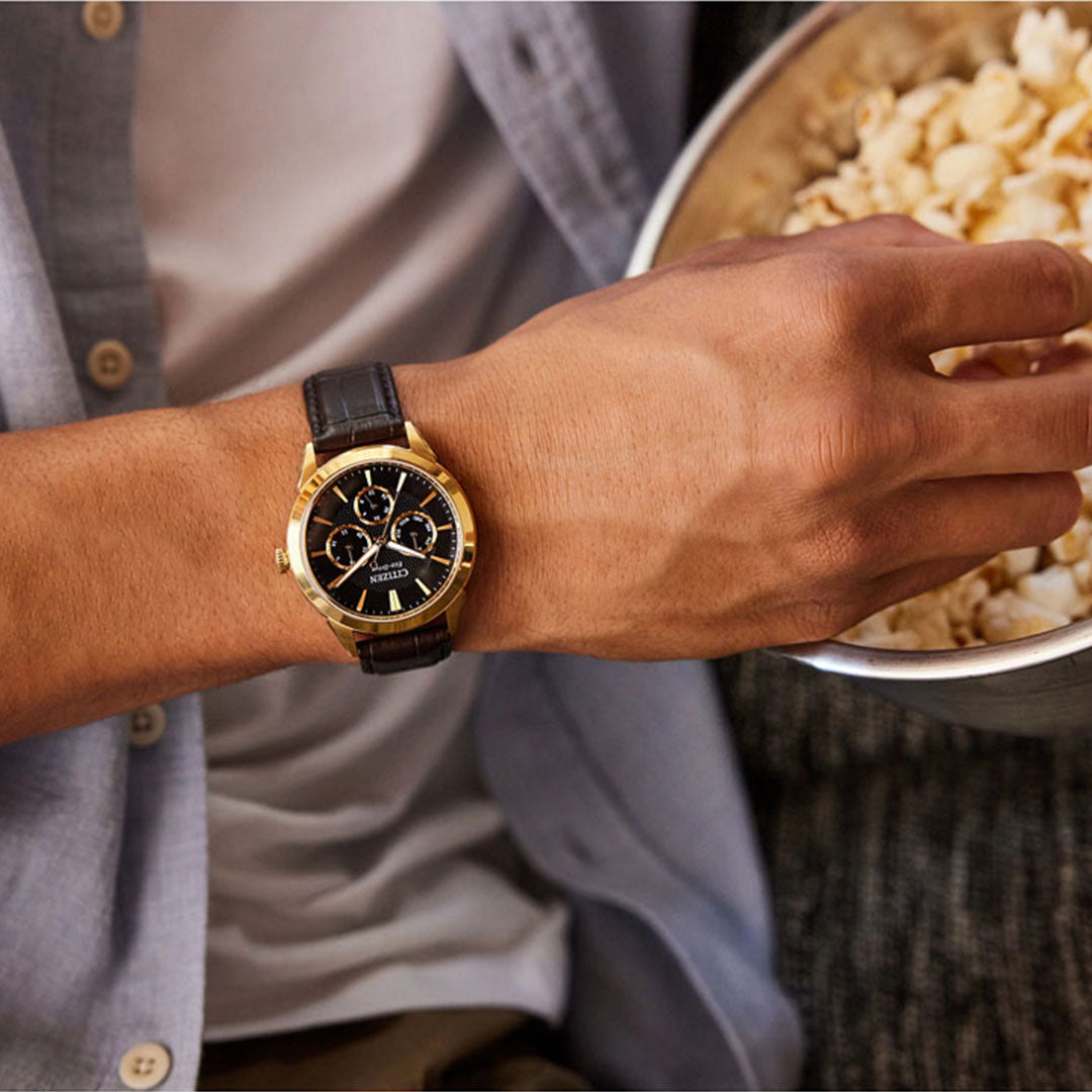 An individual wearing a sophisticated CITIZEN Rolan Eco-Drive 40mm Watch, featuring a gold wristwatch with a black dial and leather strap, is holding a bowl of popcorn. The person is dressed in a light blue shirt and is sitting on a sofa.