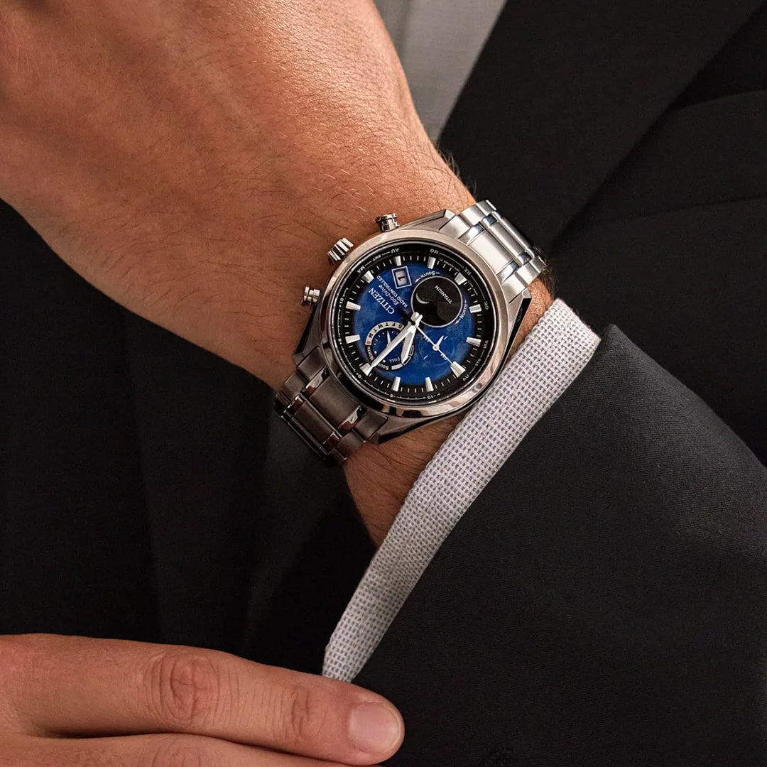 A person in a black suit jacket and white shirt wears the CITIZEN Tsuki-yomi A-T Atomic Time Keeping Super Titanium Eco-Drive 43mm Watch, featuring a silver case, blue and black dial, multiple subdials, date display, and refined craftsmanship.