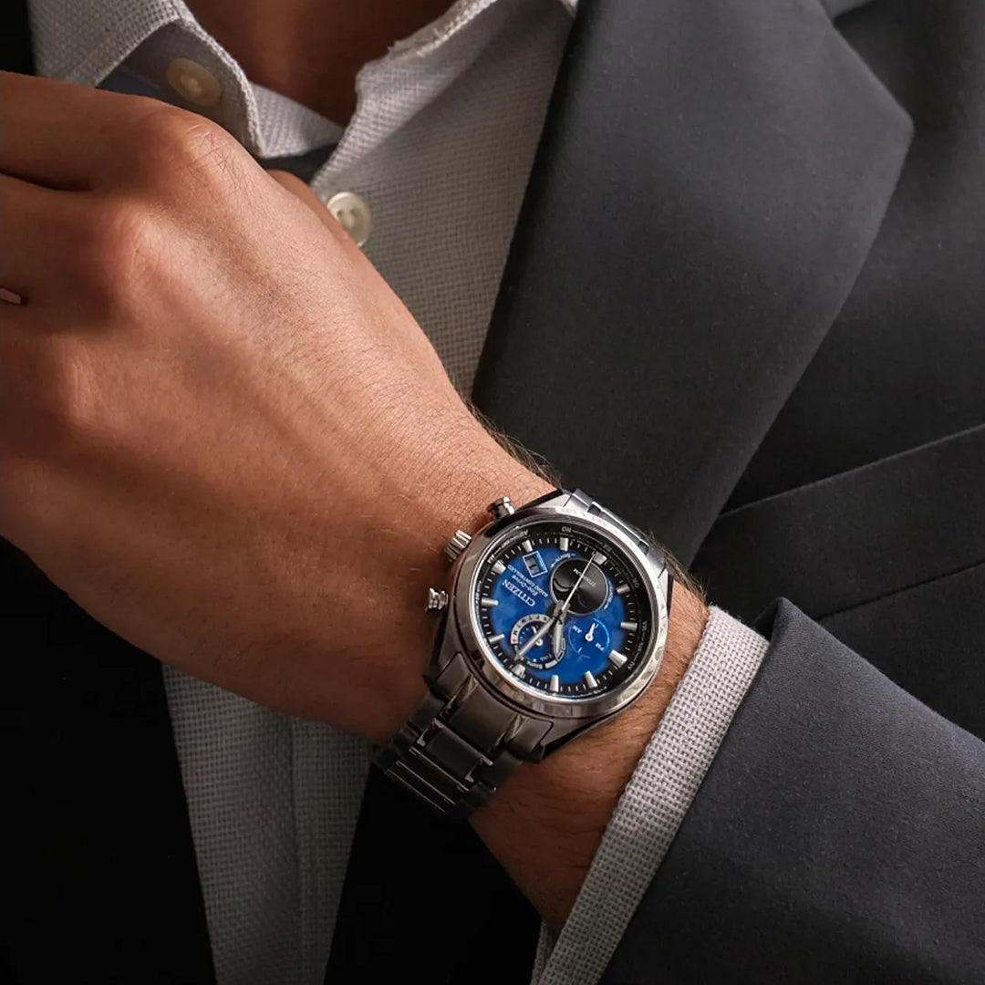 A person in a dark suit and light shirt adjusts their collar, showcasing the CITIZEN Tsuki-yomi A-T Atomic Time Keeping Super Titanium Eco-Drive 43mm Watch with a blue and black dial on their left wrist.