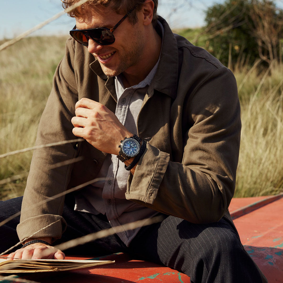 A man wearing sunglasses and a brown jacket sits outdoors on a red surface, reading a book. He's in a grassy area, smiling, with his left hand resting on his knee, displaying the CITIZEN Promaster Geo Trekker Limited Edition Eco-Drive 46mm Watch.