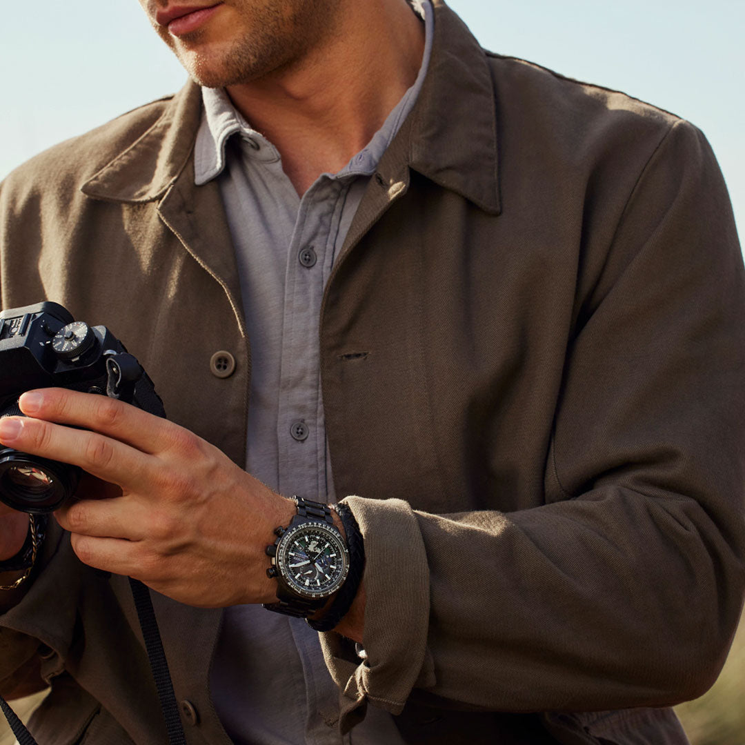 A person in a brown jacket holds a camera, their CITIZEN Promaster Geo Trekker Limited Edition Eco-Drive 46mm Watch displayed prominently. The focus is on their hands and the watch, with a hint of a grey shirt visible underneath the jacket. The background is softly blurred.