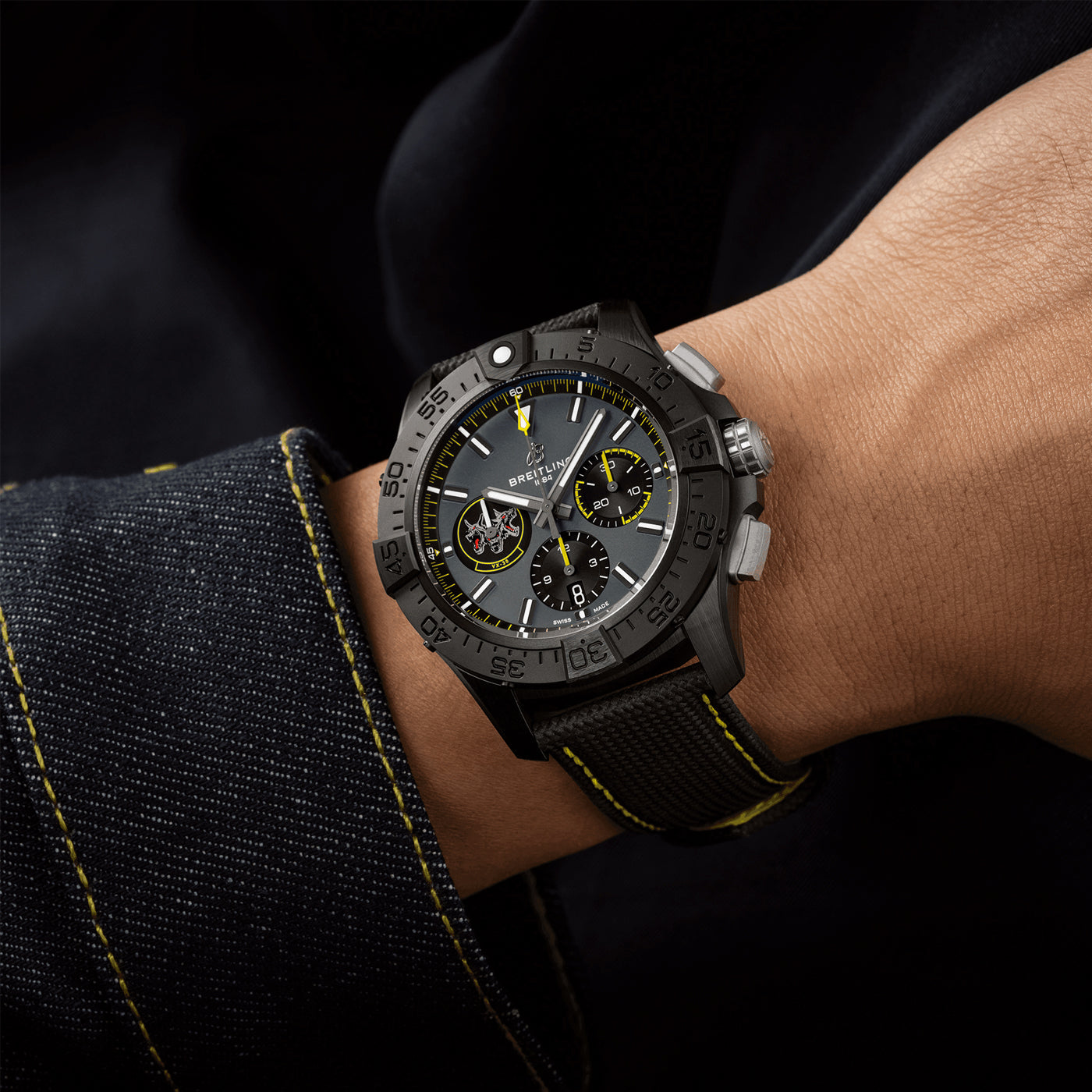 A close-up of a person's wrist showcases the BREITLING Avenger B01 Chronograph 44 Night Mission USN Bloodhounds, a rugged black watch akin to those favored by U.S. Navy jet pilots. It features a large face with multiple dials and yellow accents, while its textured strap complements the dark long-sleeved garment with visible stitching.