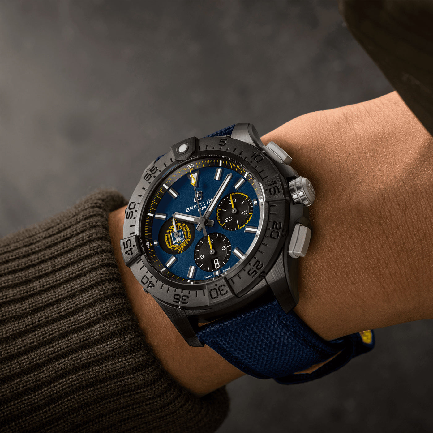 A person's wrist adorned with a Breitling Avenger B01 Chronograph 44 Night Mission US Naval Academy watch, showcasing a striking blue dial, black bezel, and a blue textured strap. The distinguished timepiece from Breitling features three subdials and a date display, all encased in ceramic. The individual is dressed in a brown sweater against a softly blurred background.