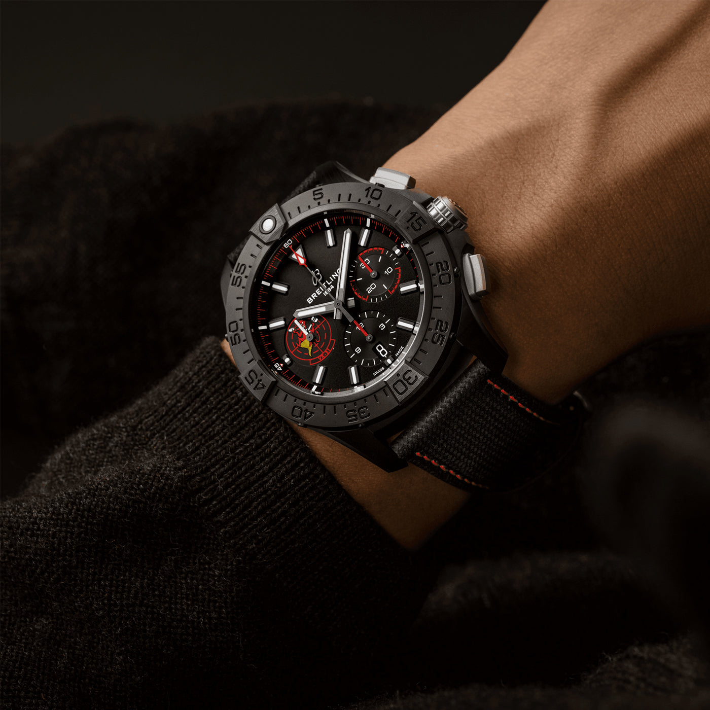 A close-up of a wrist wearing the BREITLING Avenger B01 Chronograph 44 Night Mission USN Dust Devils watch, featuring a textured band and red accents on the dial. With its bold design, large bezel, and prominent hour markers, it captures the spirit of Air Test Squadrons. The wearer pairs it with a dark, knitted sweater.