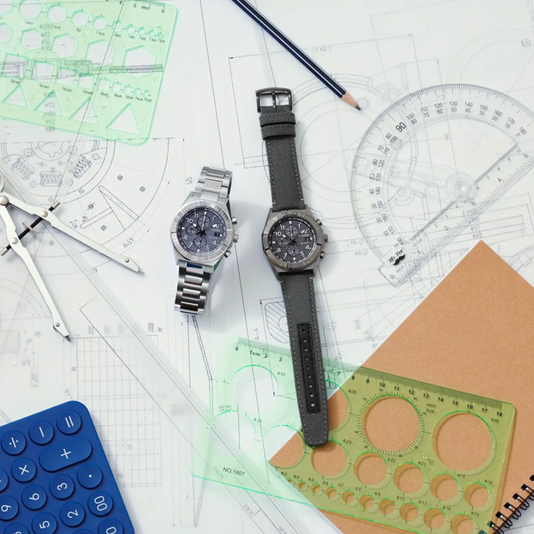 The CITIZEN Nighthawk Eco-Drive 42mm Watch by CITIZEN rests on technical drawings alongside another wristwatch, surrounded by drafting tools including green templates, a pencil, a blue calculator, a compass, a protractor, and a brown notebook.
