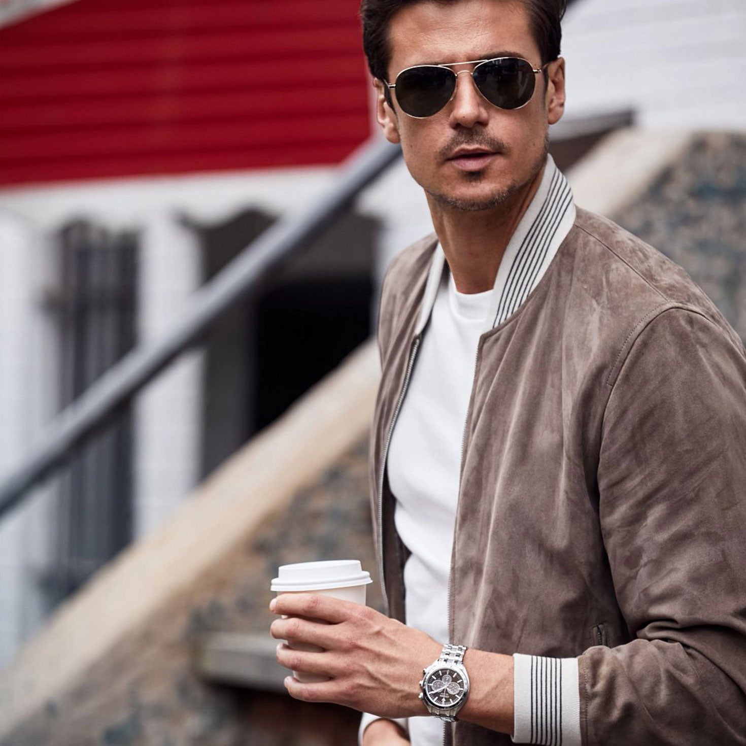 A man wearing sunglasses, a brown jacket, and a white shirt holds a coffee cup. He stands outdoors against an urban backdrop with stairs. The CITIZEN Carson Eco-Drive 43mm Watch adorns his left wrist, completing his modern look.