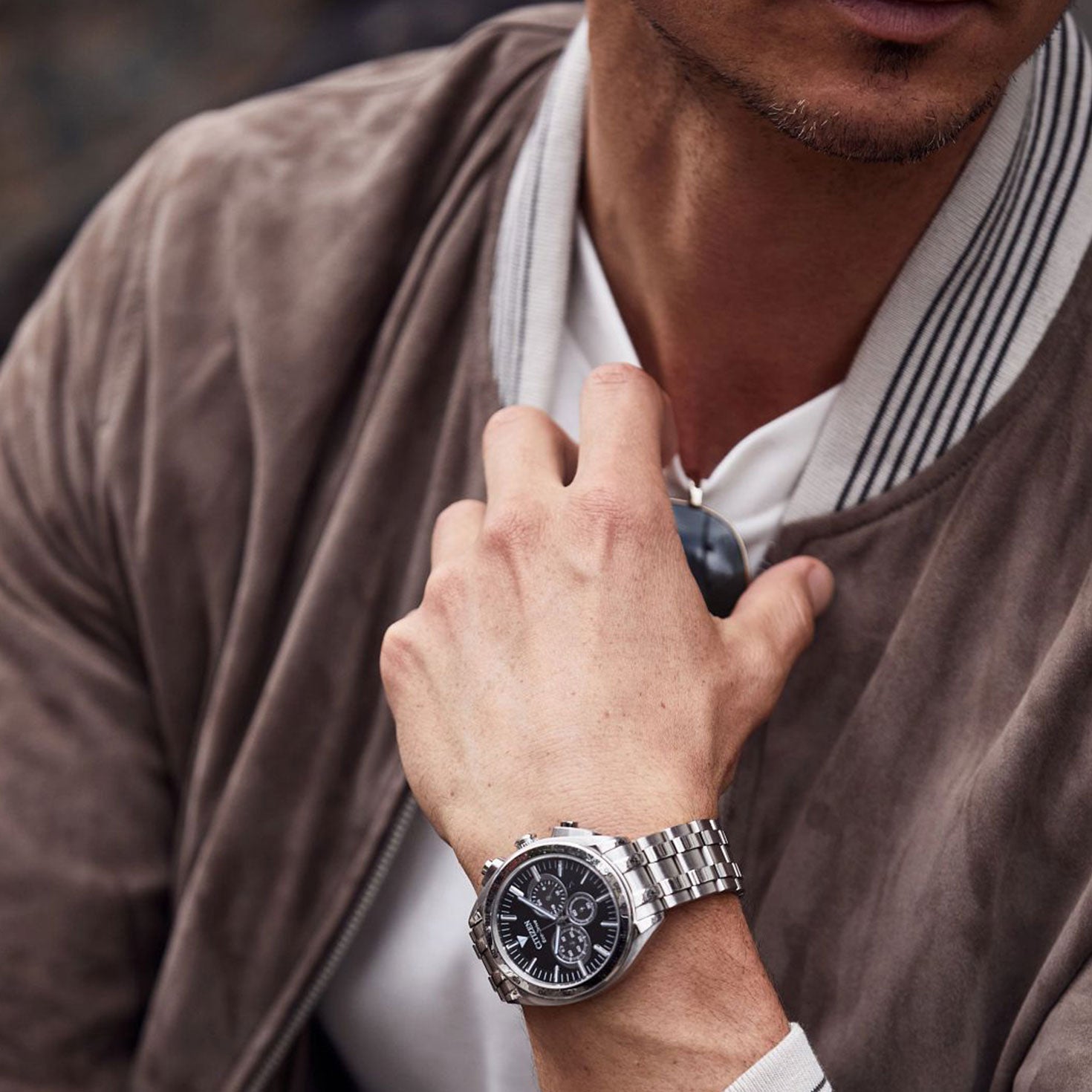 A person in a brown suede jacket and white shirt holds sunglasses, their wrist adorned with the CITIZEN Carson Eco-Drive 43mm watch, a sleek silver chronograph timepiece featuring a detailed black face.