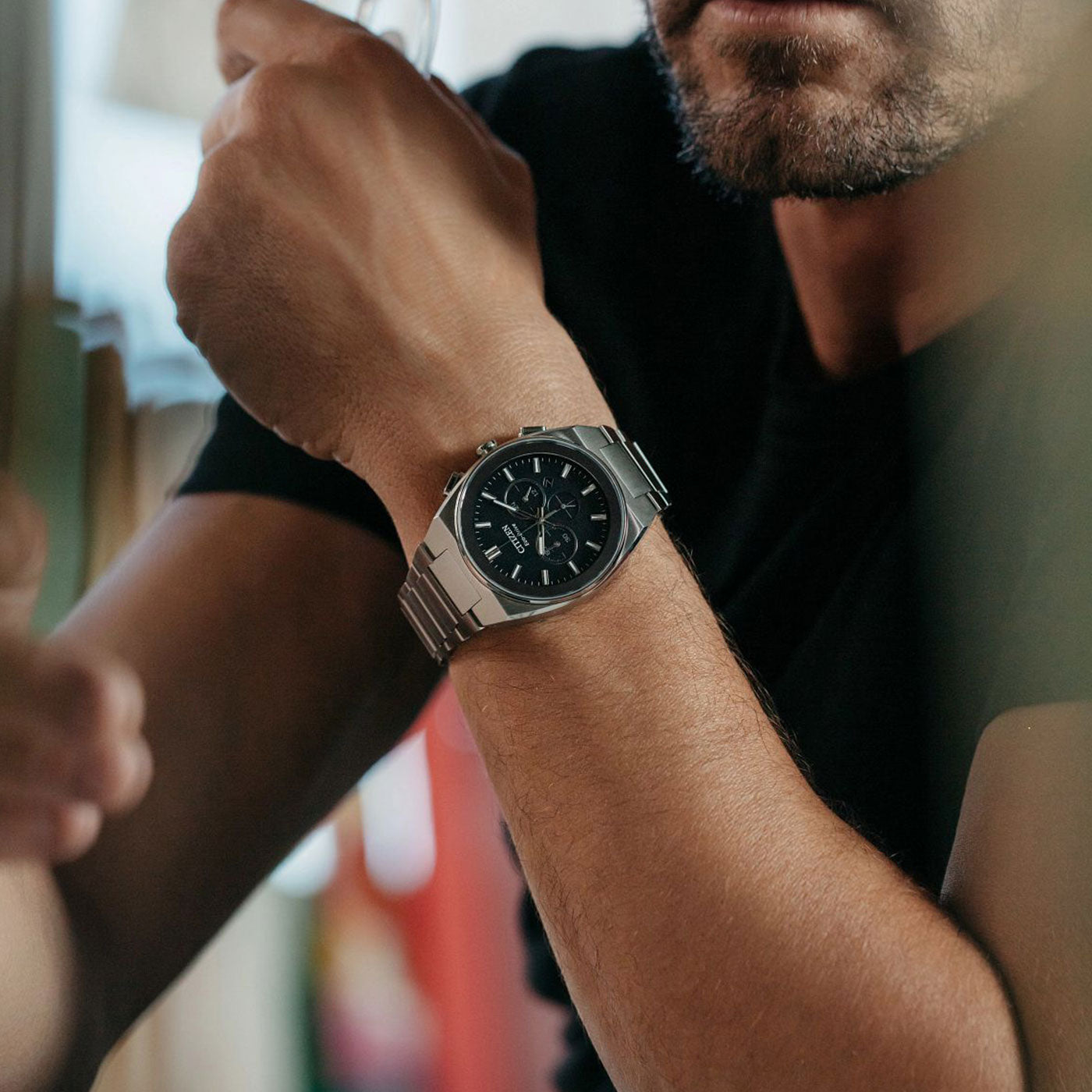 A person wearing the CITIZEN Axiom SC Eco-Drive 43mm watch, featuring a sleek metal design with a dark face and a silver band, holds a drink. The focus is on this stylish CITIZEN watch, highlighting its modern design. Casually dressed in a black shirt, they exude effortless elegance.