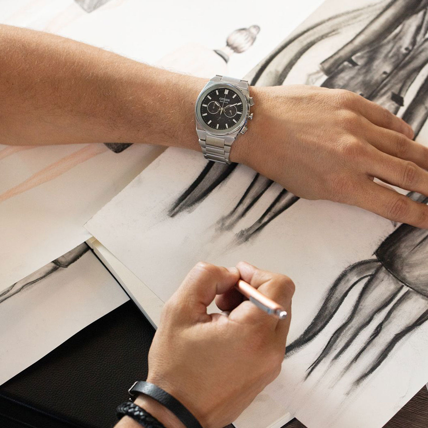 While two hands sketch on paper, one dons the sleek CITIZEN Axiom SC Eco-Drive 43mm Watch, and the other wields a pencil. Abstract shapes and lines come alive on the page, inspired by organic forms. The scene highlights the creativity and precision of this artistic journey, capturing movement and imagination in every stroke.