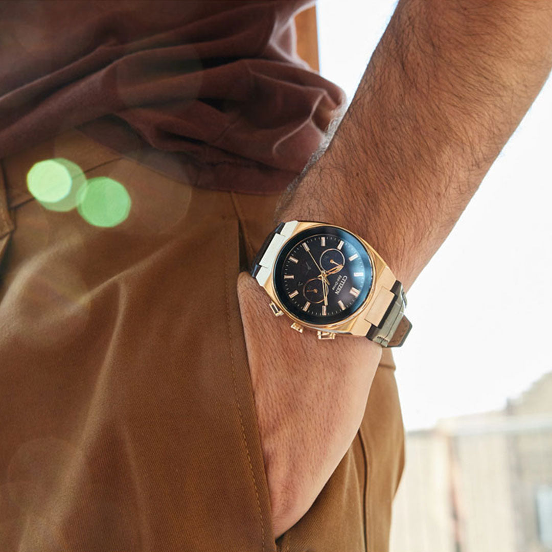 A person dressed in a brown shirt and pants has their hand in their pocket, showcasing a CITIZEN Axiom SC B620 Eco-Drive 43mm Watch, featuring a rose gold-tone with a black face and metallic band. Sunlight generates lens flare effects in the photograph.