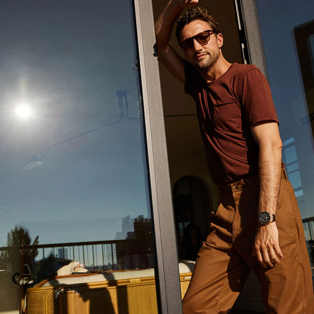 A man in a brown t-shirt and pants leans against a glass door, wearing sunglasses and the CITIZEN Axiom SC B620 Eco-Drive 43mm Watch. The scene reflects a sunny day with a cityscape visible in the window's reflection, capturing the essence of his Eco-Drive technology timepiece.