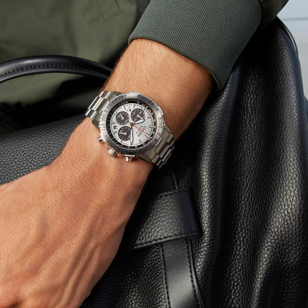 A person wearing the CITIZEN Promaster Navihawk Eco-Drive 40mm Watch, featuring a silver stainless steel finish and a black-and-white dial with subdials, rests their wrist on a black leather bag while donning a green long-sleeve shirt, illustrating an elegant blend of style and functionality.