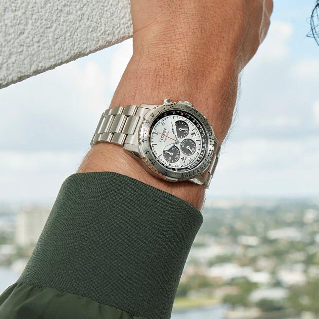 A close-up reveals the timeless elegance of a CITIZEN Promaster Navihawk Eco-Drive 40mm Watch on a wrist, its stainless steel design featuring a white face and three black sub-dials. The wearer dons a green long-sleeve shirt against a blurred cityscape under cloudy skies.