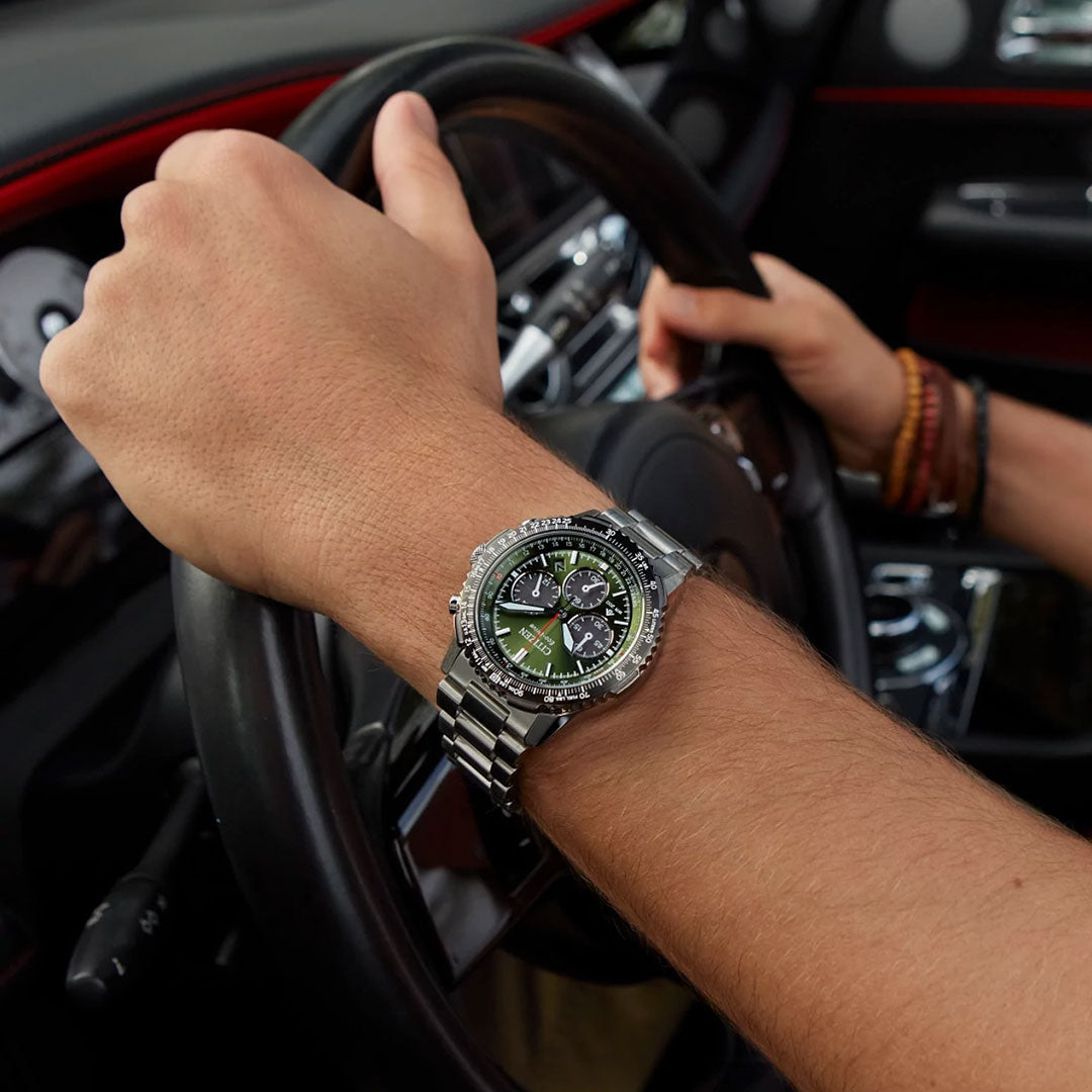 A person wearing a CITIZEN Promaster Navihawk Eco-Drive 40mm watch with a stainless steel bracelet drives a car, one hand on the wheel and dashboard controls visible in the background.