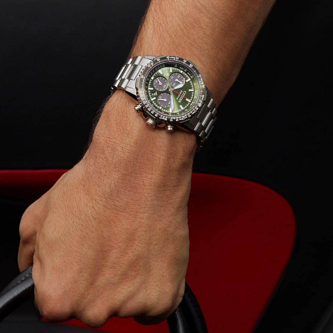A wrist sports the CITIZEN Promaster Navihawk Eco-Drive 40mm Watch, featuring a silver chronograph design and green face, while gripping a black steering wheel against a dark background accented with red.