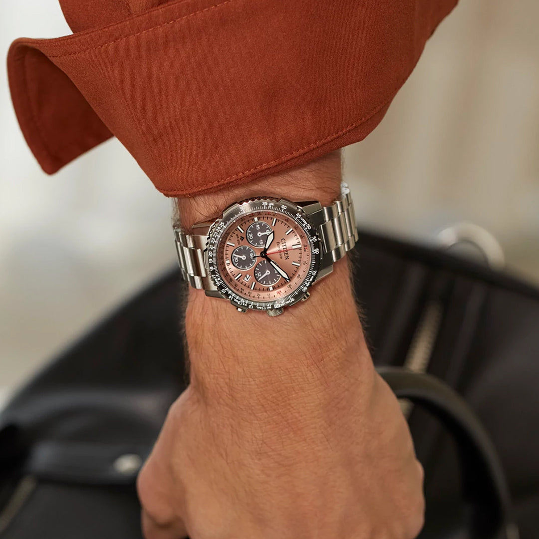 A person wearing a CITIZEN Promaster Navihawk Eco-Drive 40mm watch, with a pink face and three subdials. The sleeve of an orange-brown jacket appears as the hand rests on a black leather object. The watch features a silver bezel and stainless steel band.