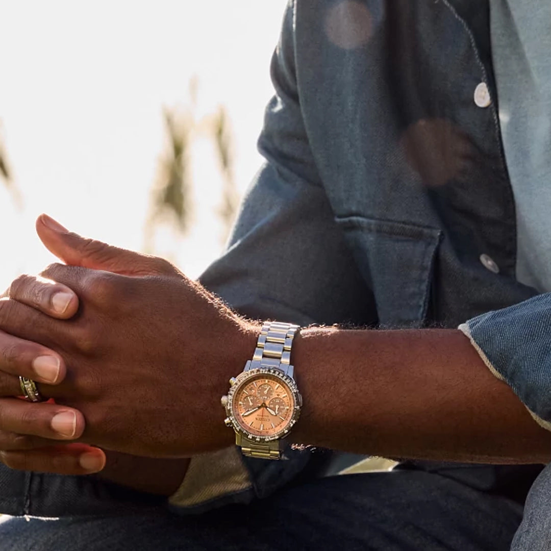 A person in a denim jacket sits with folded hands, showcasing the CITIZEN Promaster Navihawk Eco-Drive 40mm Watch with a copper face and stainless steel case. Sunlight softly highlights its Eco-Drive technology.