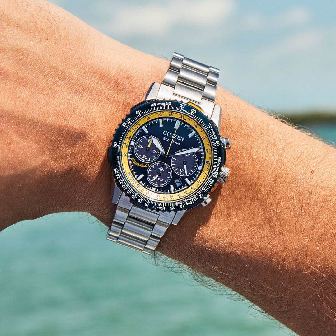 A person wearing the CITIZEN Promaster Navihawk Eco-Drive 40mm Watch stands by the water, sunlight gleaming off its stainless steel band.