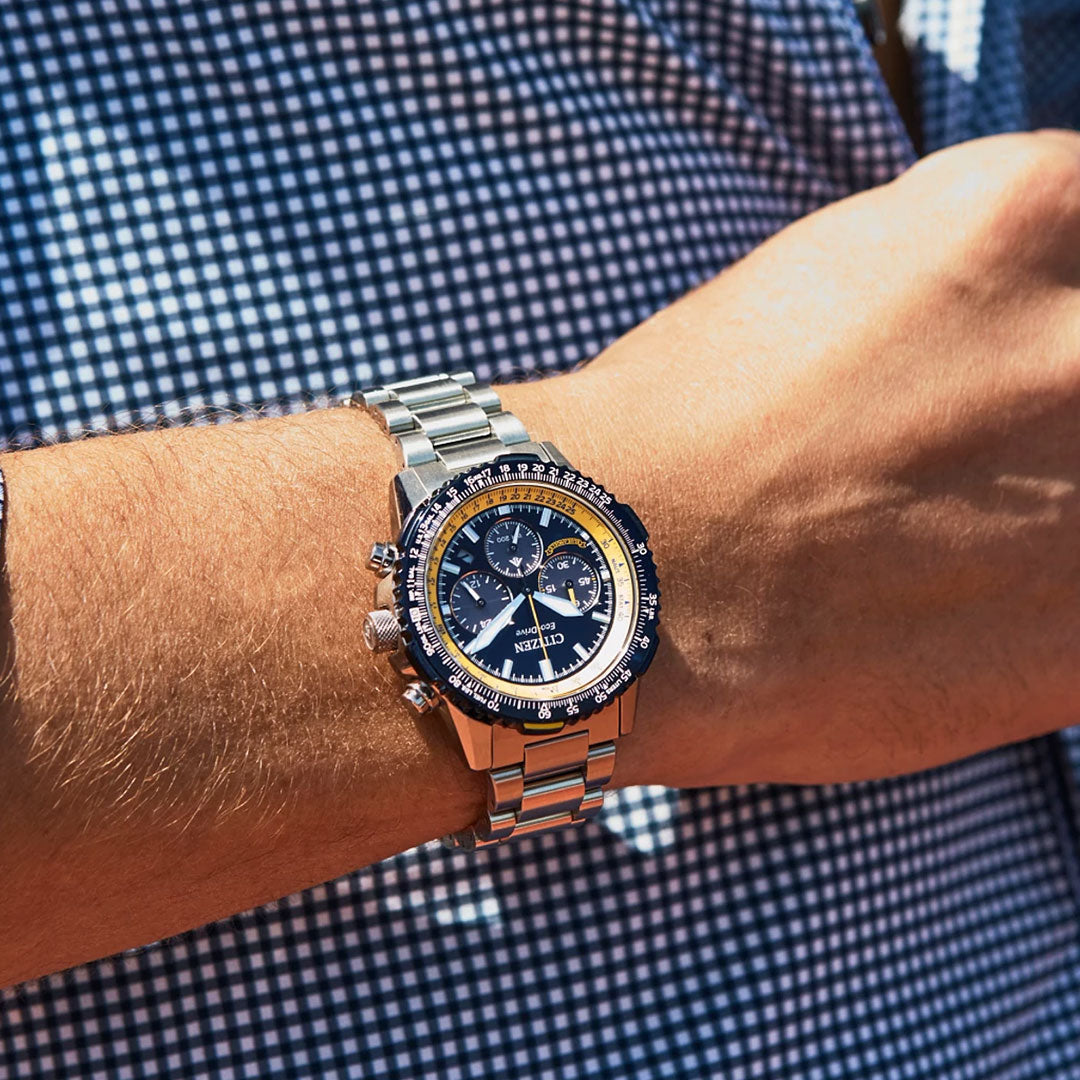 A close-up shows a person wearing the CITIZEN Promaster Navihawk Eco-Drive 40mm Watch with a black and yellow dial, silver stainless steel band, and multiple subdials—standing out against a blue and white checkered shirt.