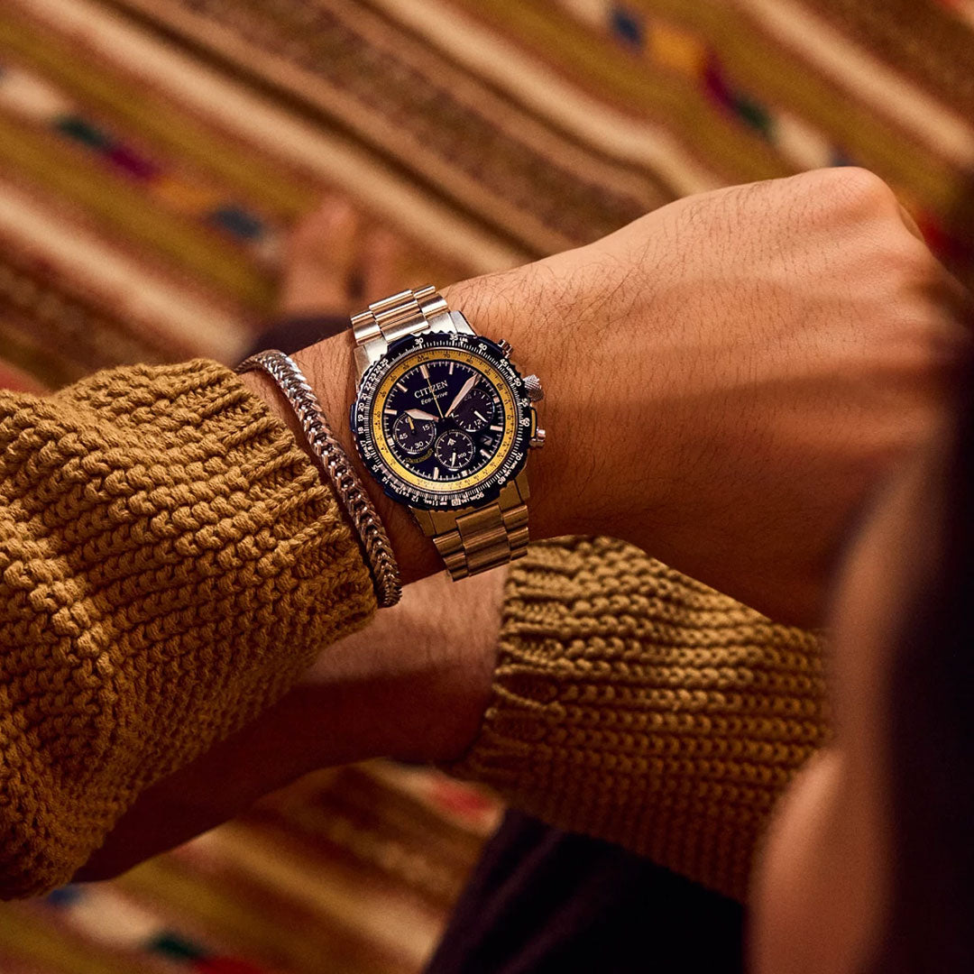 Wearing a chunky mustard sweater and silver bracelet, someone checks their CITIZEN Promaster Navihawk Eco-Drive 40mm Watch as its dark dial shines. A colorful patterned rug in the background adds a vibrant accent.