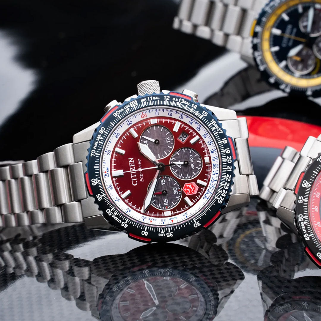 A CITIZEN Promaster Navihawk Eco-Drive 40mm stainless steel watch with a red and white dial and silver bracelet is displayed on a reflective carbon fiber surface, with other CITIZEN watches in the background.