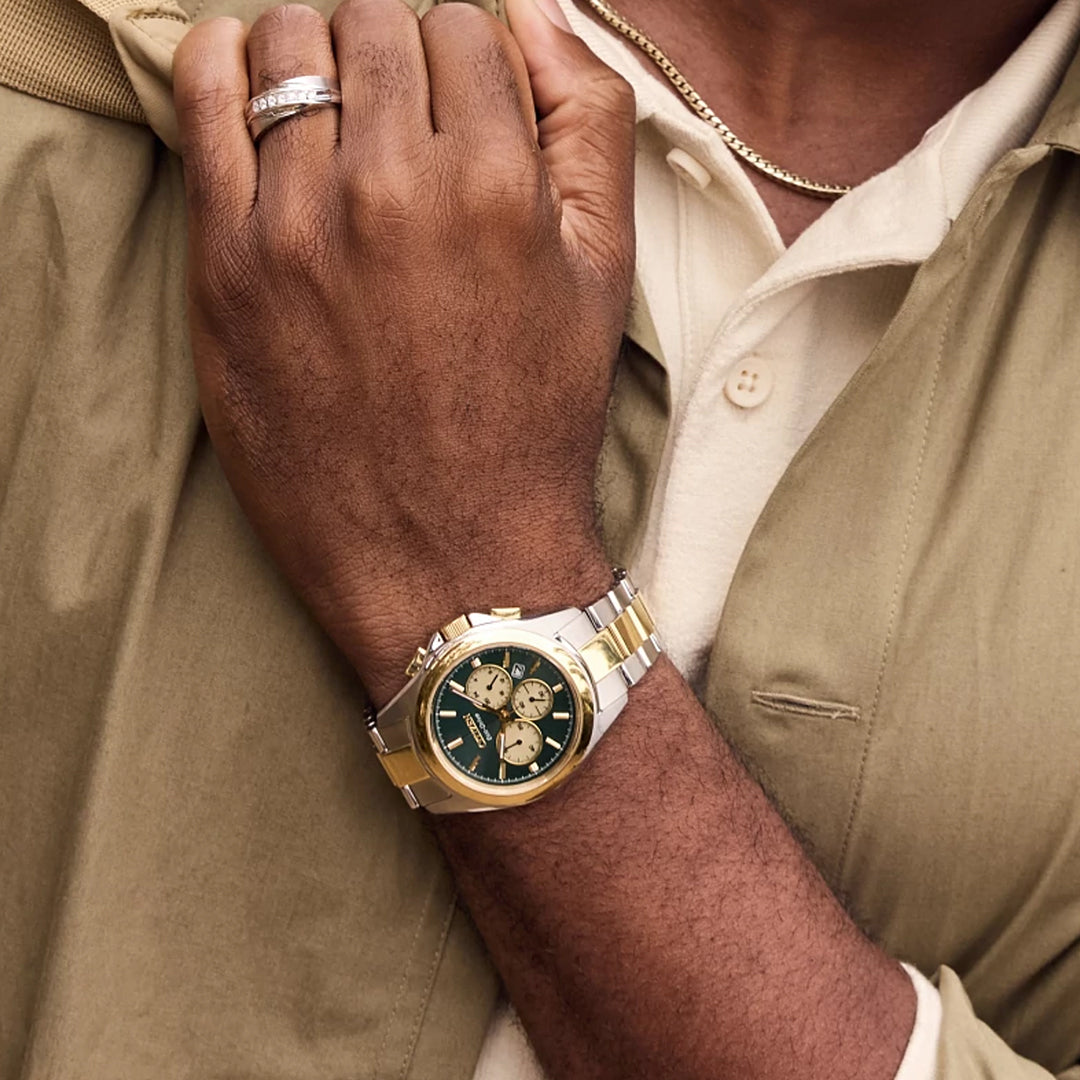 A person wearing a CITIZEN Sport Luxury Eco-Drive 40.5mm watch with a green and gold face, paired with a gold chain, beige shirt, and khaki jacket draped over their shoulder, holds their wrist.