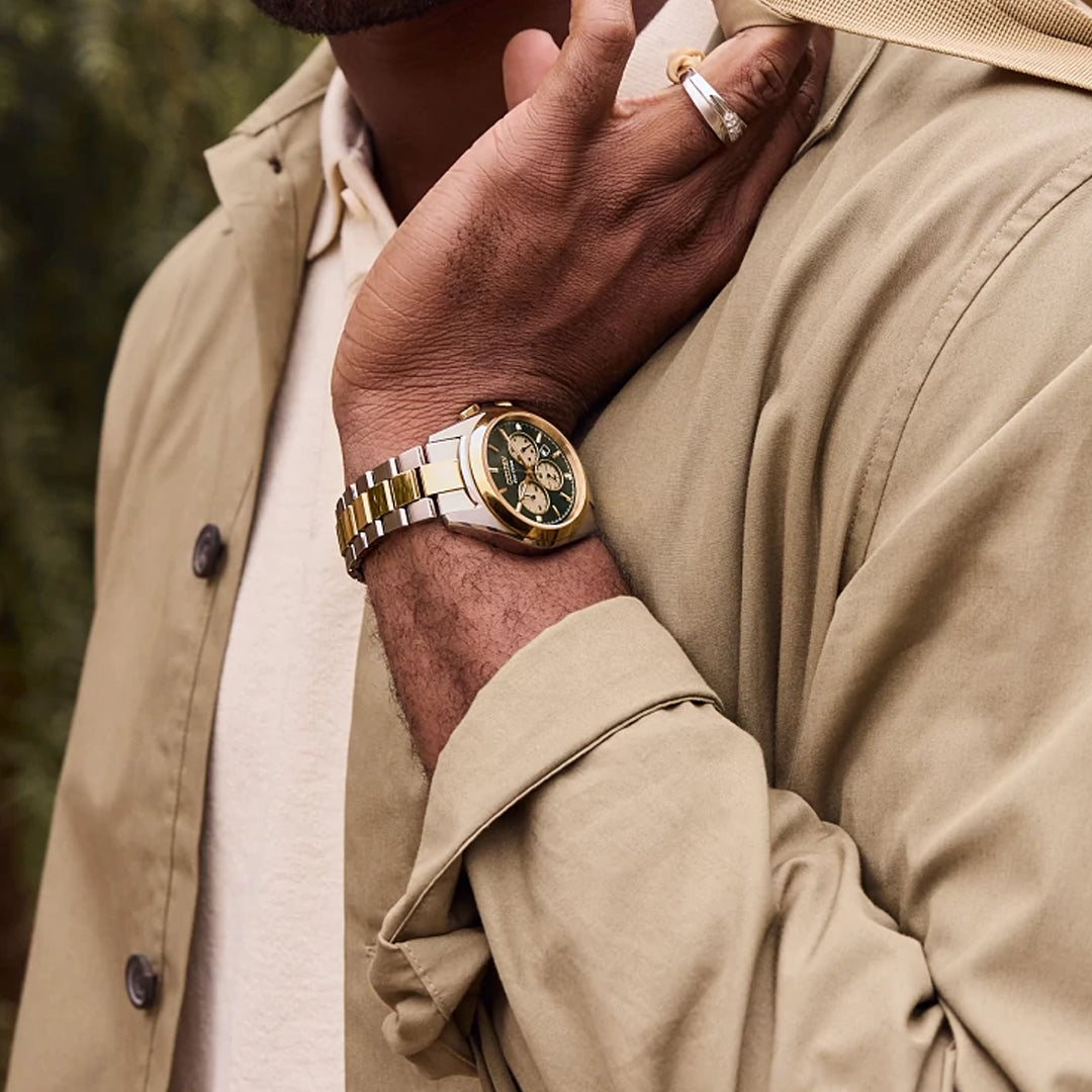 A person in a beige jacket and white shirt touches their neck, showcasing the CITIZEN Sport Luxury Eco-Drive 40.5mm Watch with a gold case and dark face. The stainless steel bracelet glints subtly, complemented by a ring, drawing attention to the watch and elegant attire against the blurred background.