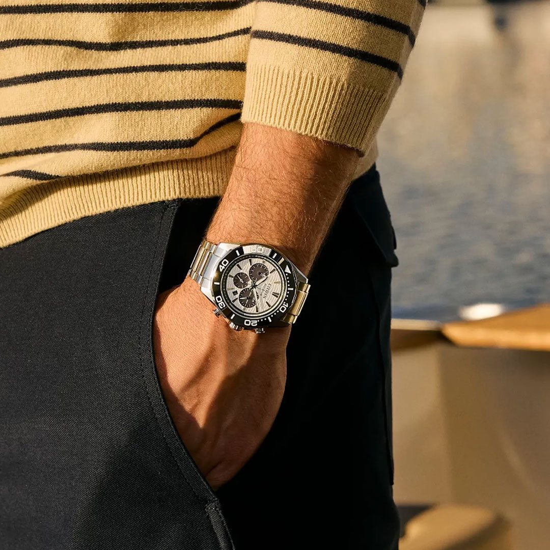 Wearing a beige and black striped sweater and dark pants, a person poses with one hand in their pocket, showcasing the CITIZEN Endeavor Chrono Eco-Drive 43mm Watch; a blurred calm body of water is visible in the background.