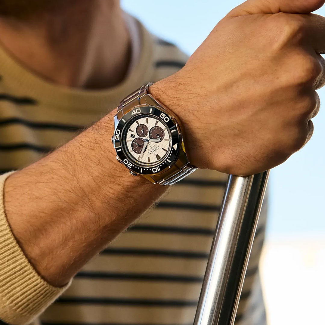 A person in a tan and black striped sweater displays the CITIZEN Endeavor Chrono Eco-Drive 43mm Watch, featuring a stainless steel case, black bezel, and light dial from CITIZEN, while holding a metal pole.