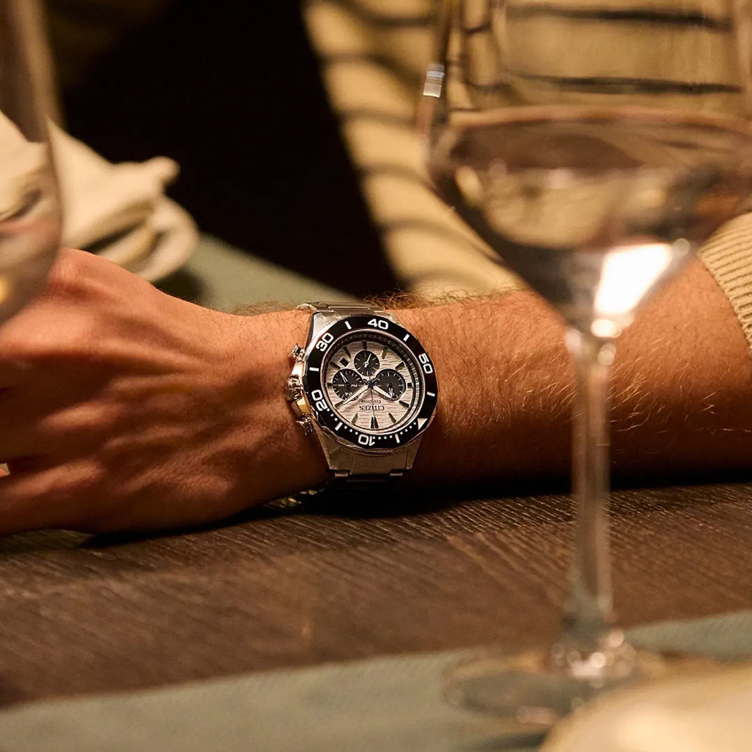 A person wearing a CITIZEN Endeavor Chrono Eco-Drive 43mm watch with a stainless steel band and black bezel rests their arm on a wooden table, surrounded by wine glasses and soft lighting.