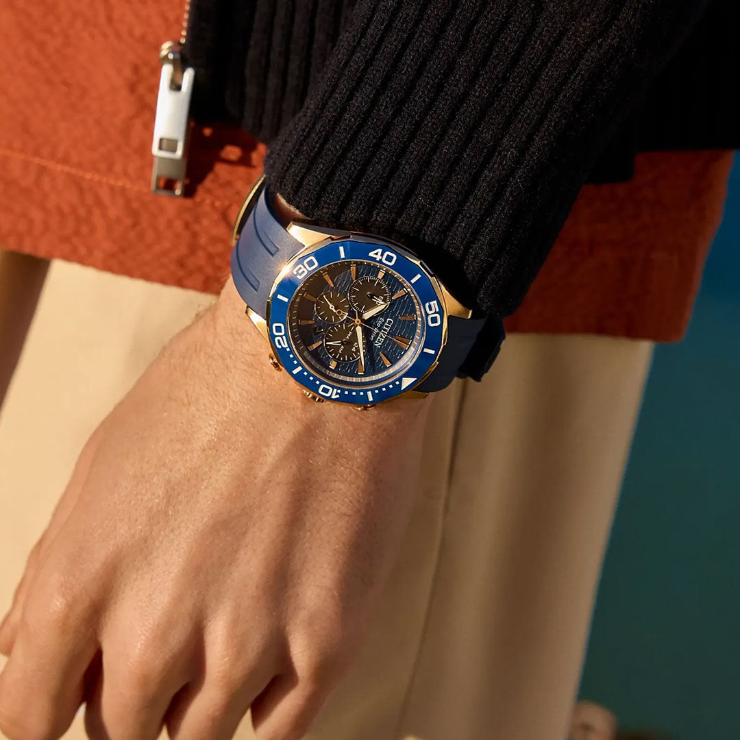 A person in a dark sweater and beige pants displays the CITIZEN Endeavor Chrono Eco-Drive 43mm Watch, featuring a blue strap, multi-dial face, and rose gold-tone stainless steel details against a softly blurred backdrop.