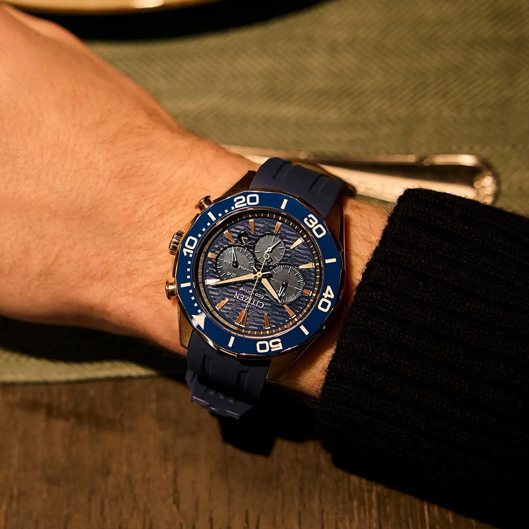 A person wearing a CITIZEN Endeavor Chrono Eco-Drive 43mm Watch with a blue rubber strap and silver accents sits at a table, with a green placemat and silverware in the background.