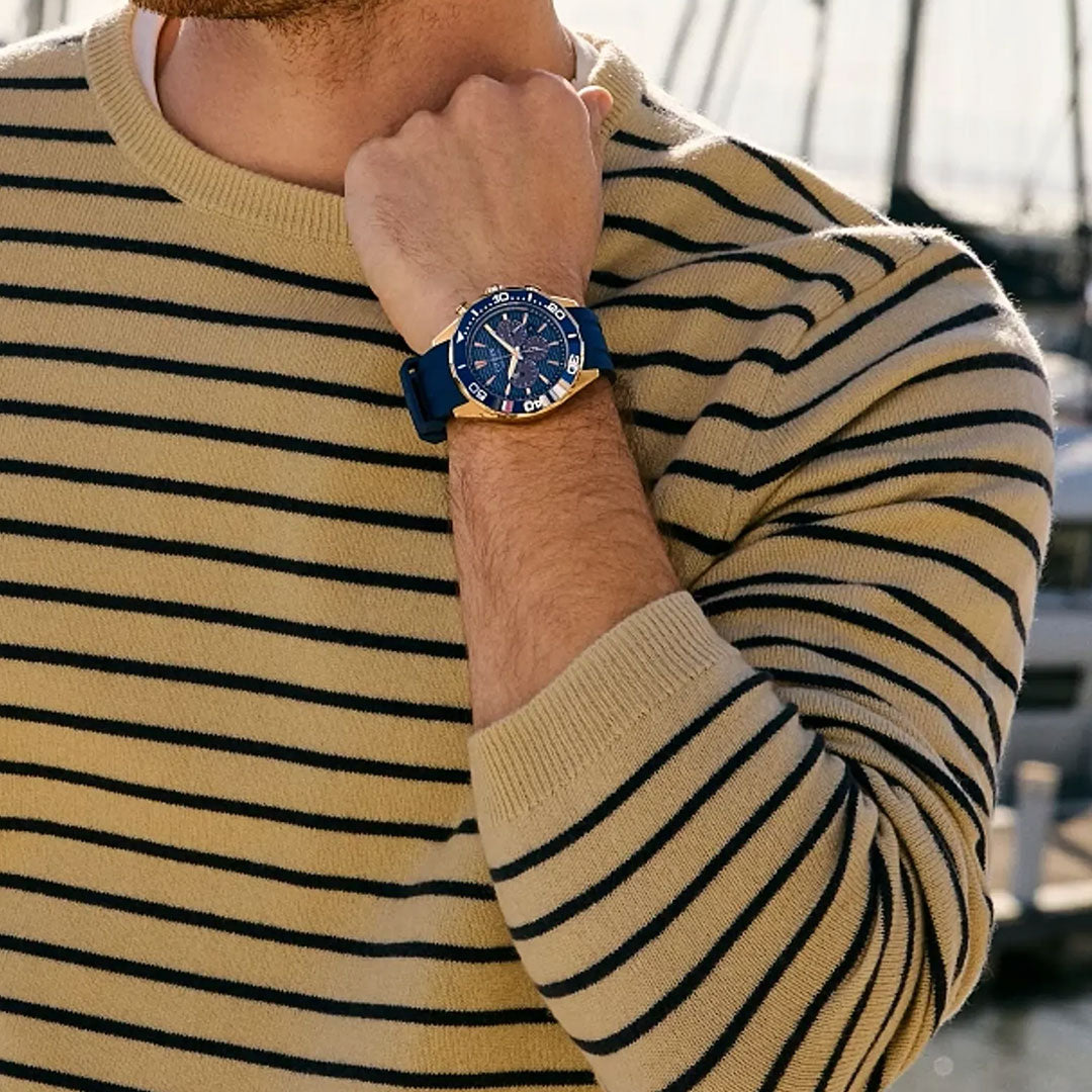 Wearing a beige and black striped sweater, a person showcases the CITIZEN Endeavor Chrono Eco-Drive 43mm Watch with blue strap and dial, standing outdoors by blurred boats, highlighting its nautical-inspired chronograph style.