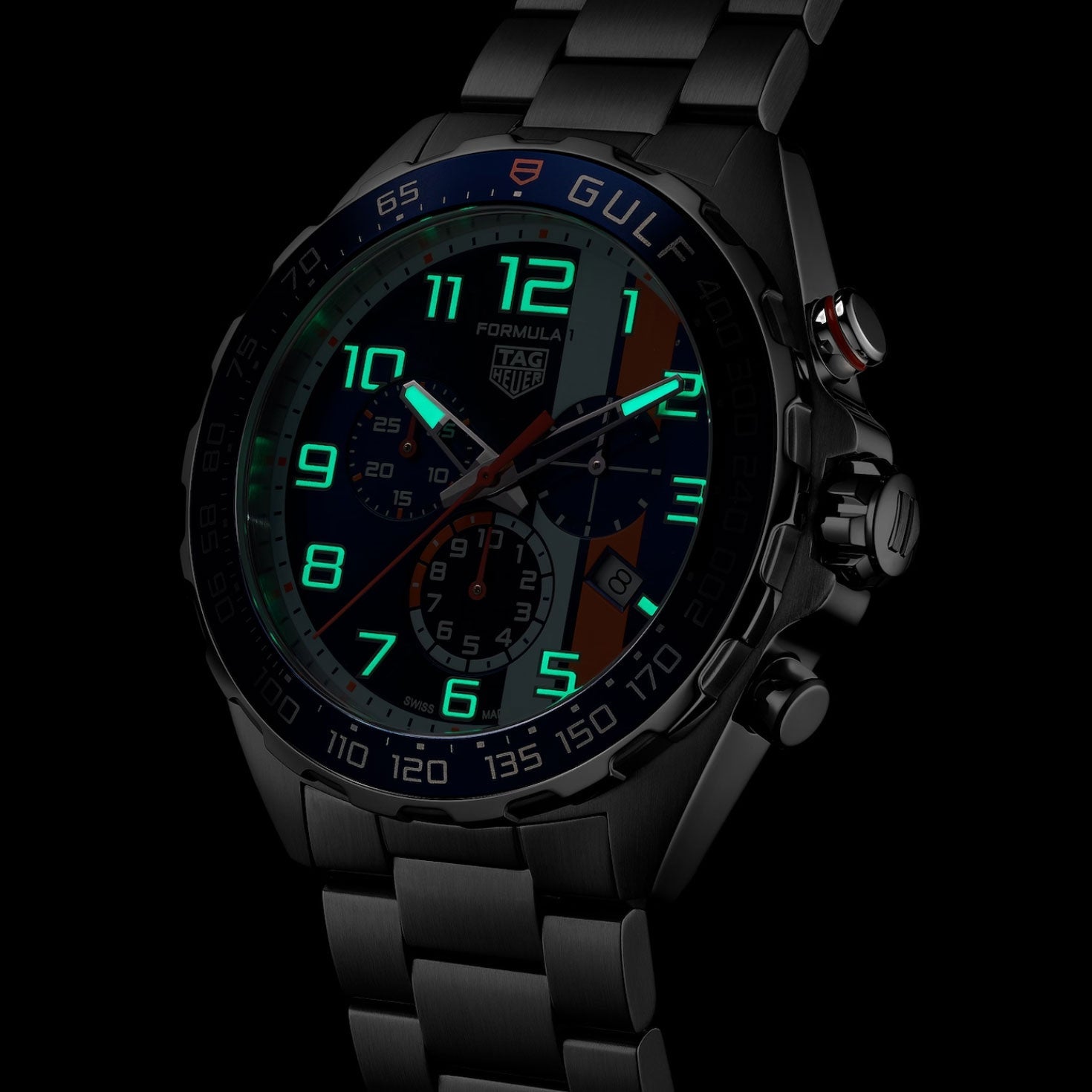 A TAG Heuer Formula 1 Chronograph x Gulf Special Edition Quartz 43mm watch in black is displayed with a sleek metal bracelet, its luminescent green numerals and hands glowing boldly in a dark setting.