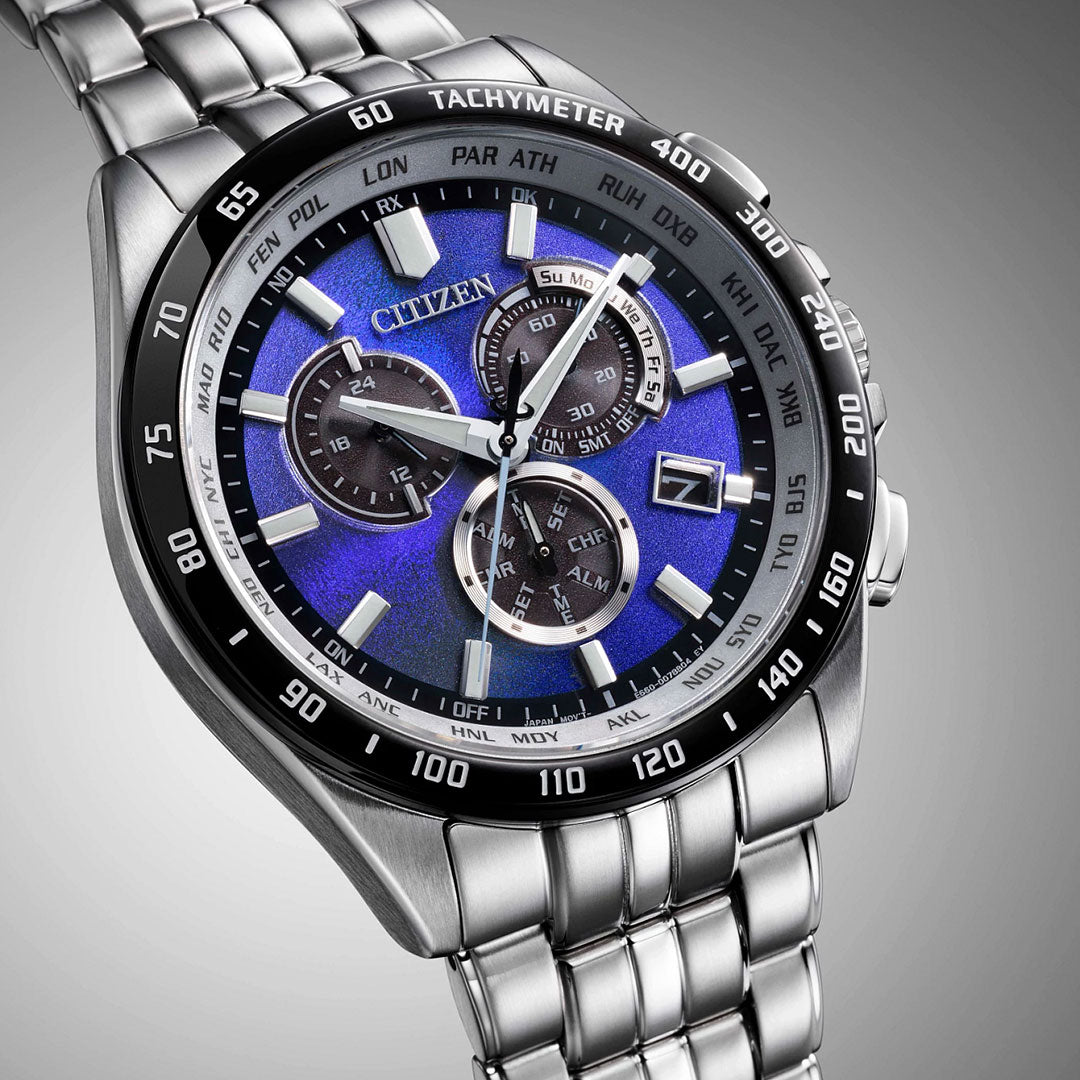 A close-up of the CITIZEN Perpetual Chrono A-T Eco-Drive 43mm watch shows its blue dial with multiple subdials, silver hour markers, and a stainless steel band set against a gradient gray background.